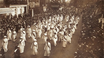 Sepia toned image of a regiment of women in uniform (white or pale clothes and a dark sash) carrying staffs and marching in formation in a suffrage parade