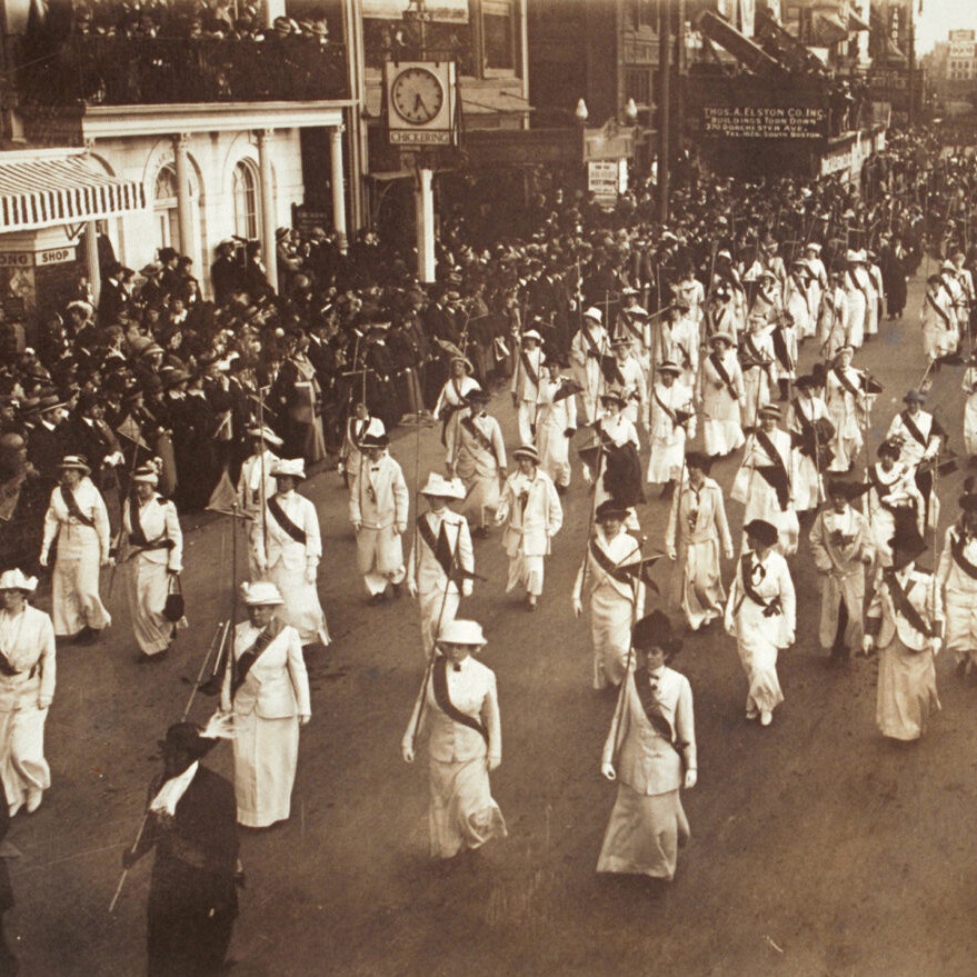Sepia toned image of a regiment of women in uniform (white or pale clothes and a dark sash) carrying staffs and marching in formation in a suffrage parade