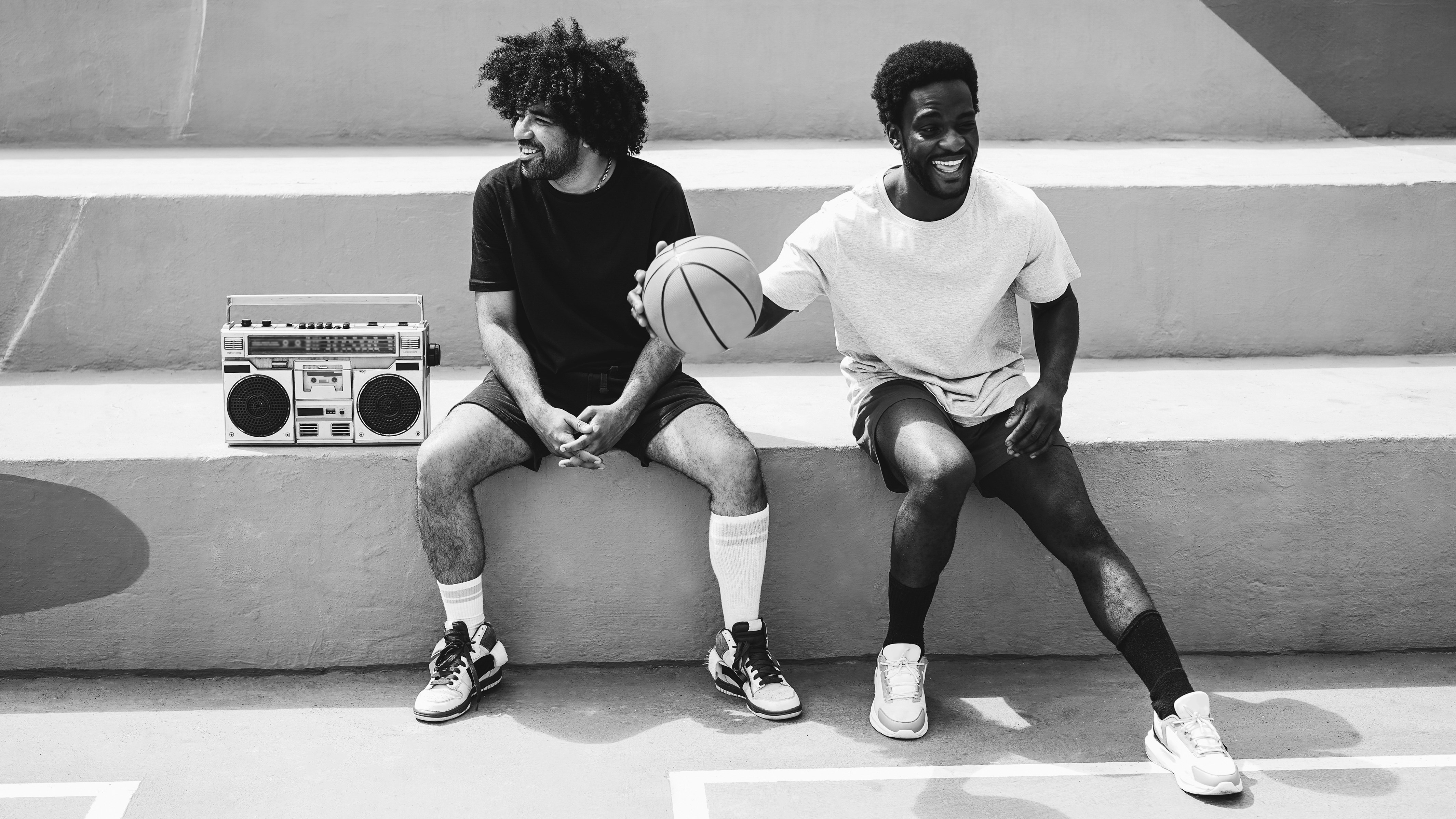 Two smiling Black men in activewear sit on concrete steps next to a boom box while one of them dribbles a basketball.