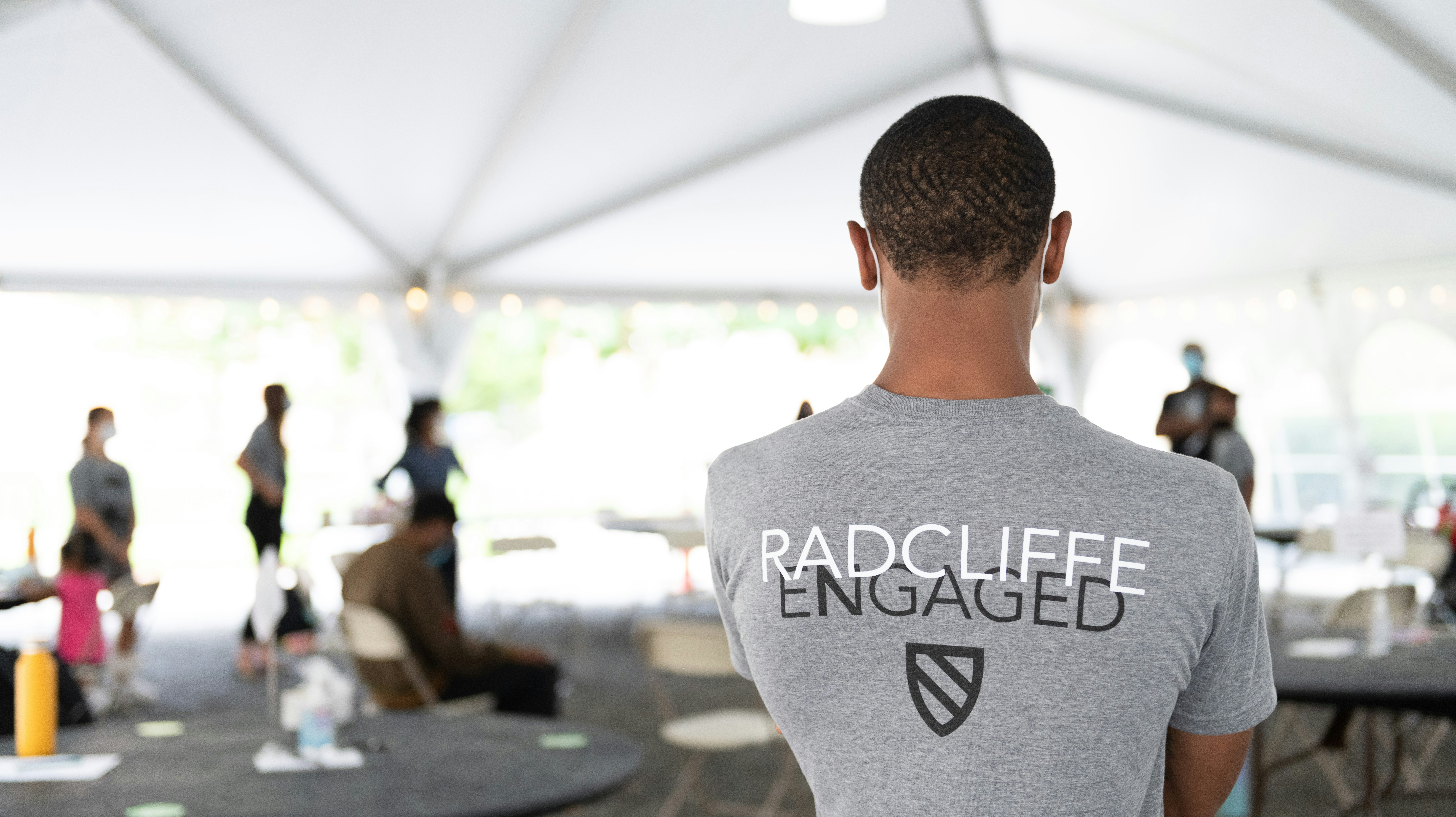 A student stands with his back to the camera in a tent in Radcliffe Yard. His t-shirt reads "Radcliffe Engaged" and displays the Radcliffe shield.
