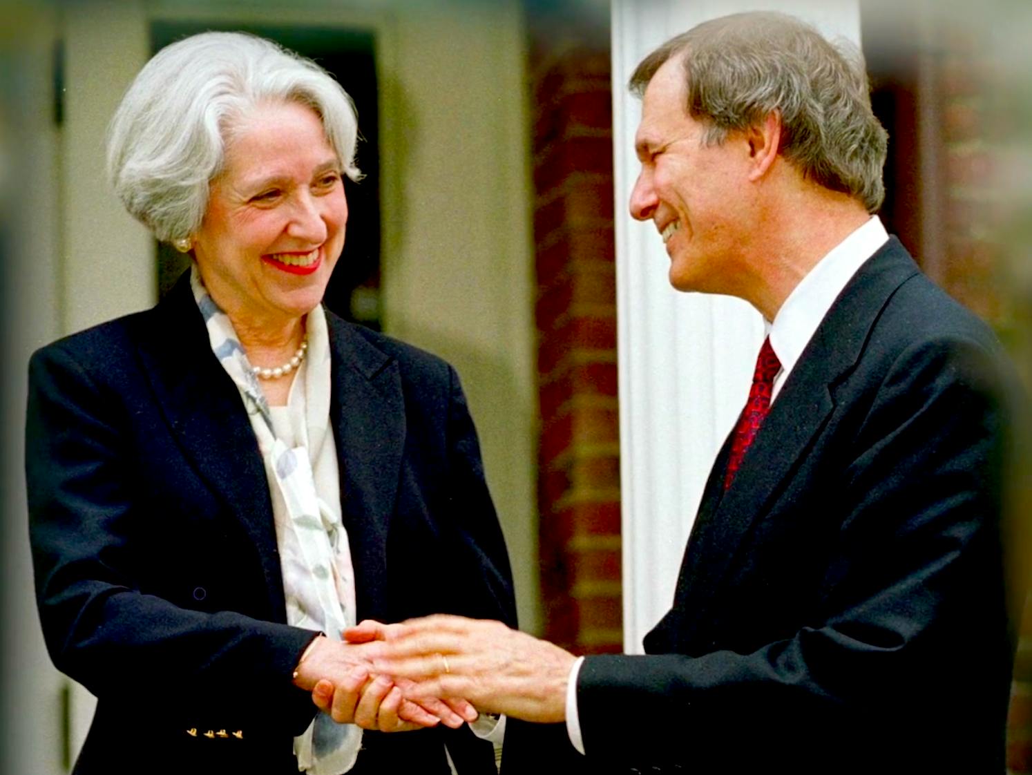 A smiling woman with white bobbed hair in a suit and pearls shakes hand with a suited man in profile.