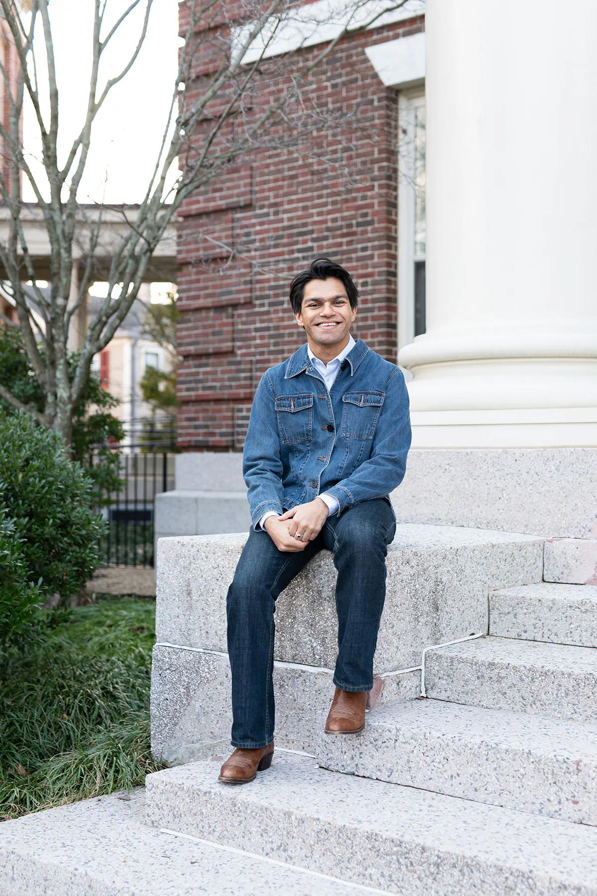 A smiling young man wearing a denim work jacket and cowboy boots sits outdoors on stone steps outside a brick building.