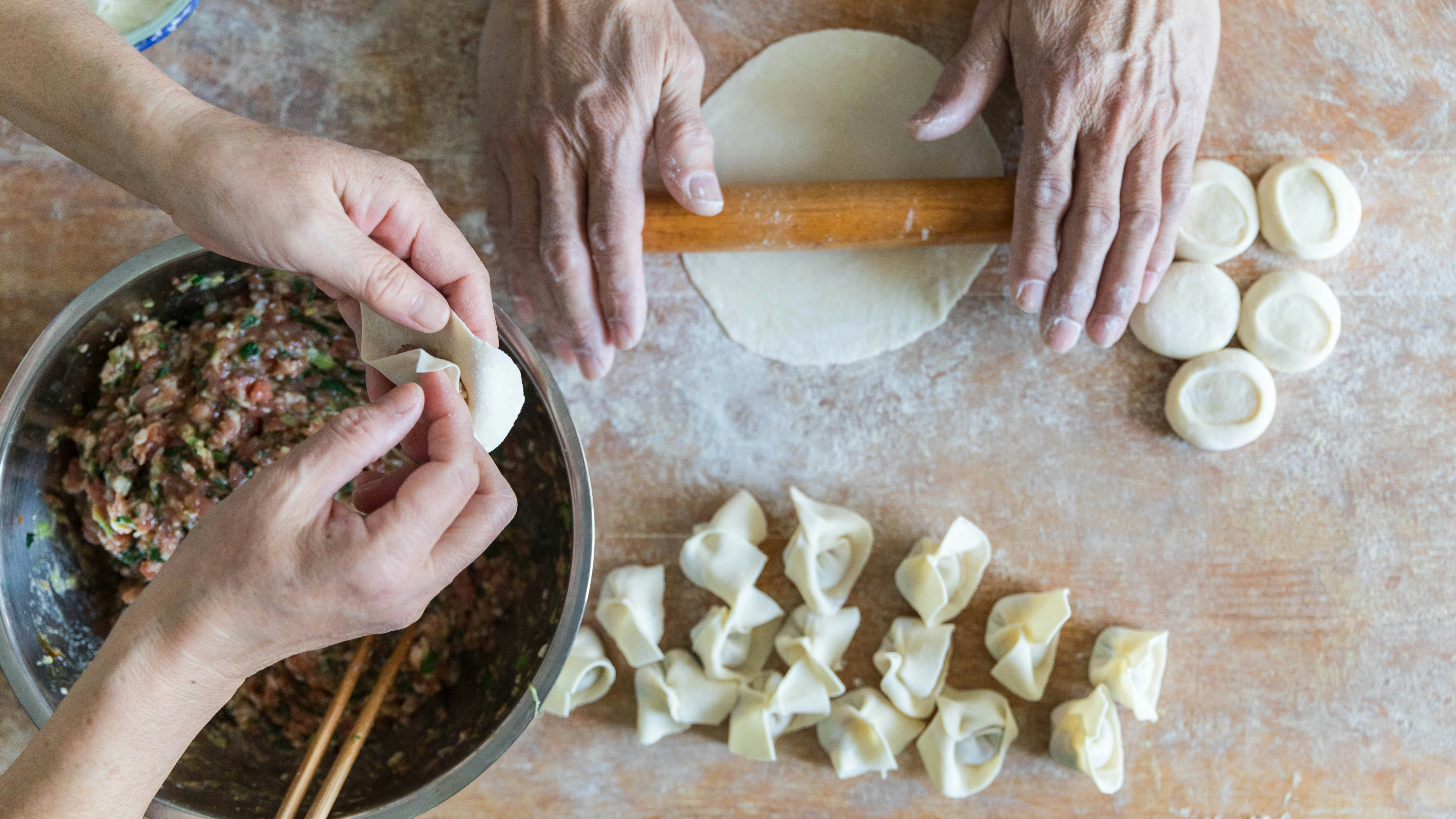 Two pairs of hands roll out and shape dumplings in a home kitchen.