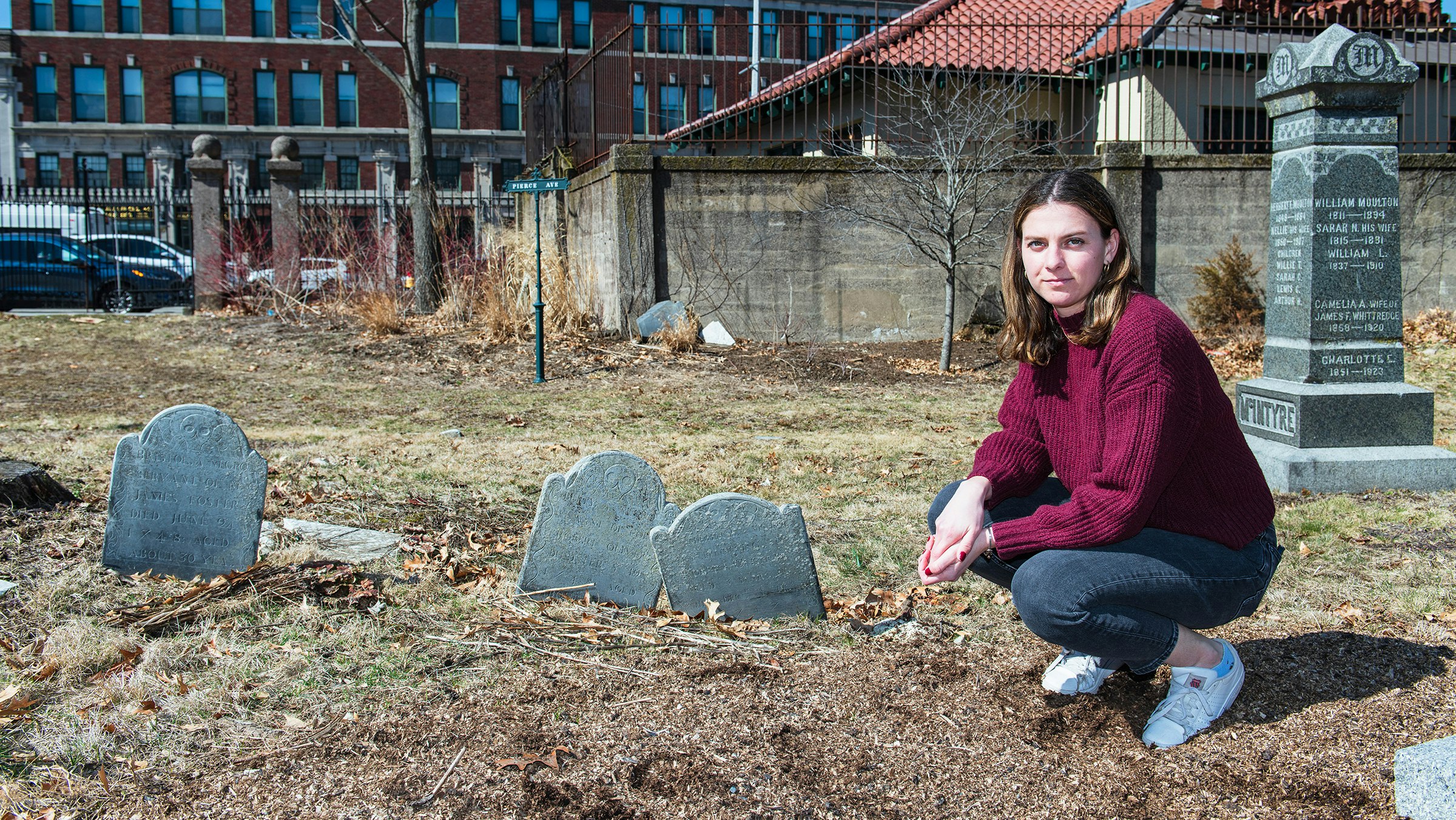 A casually dressed woman looks directly at the camera as she squats down next to modest antique gravestones situated in an urban cemetery. Low city buildings are visible in the background.