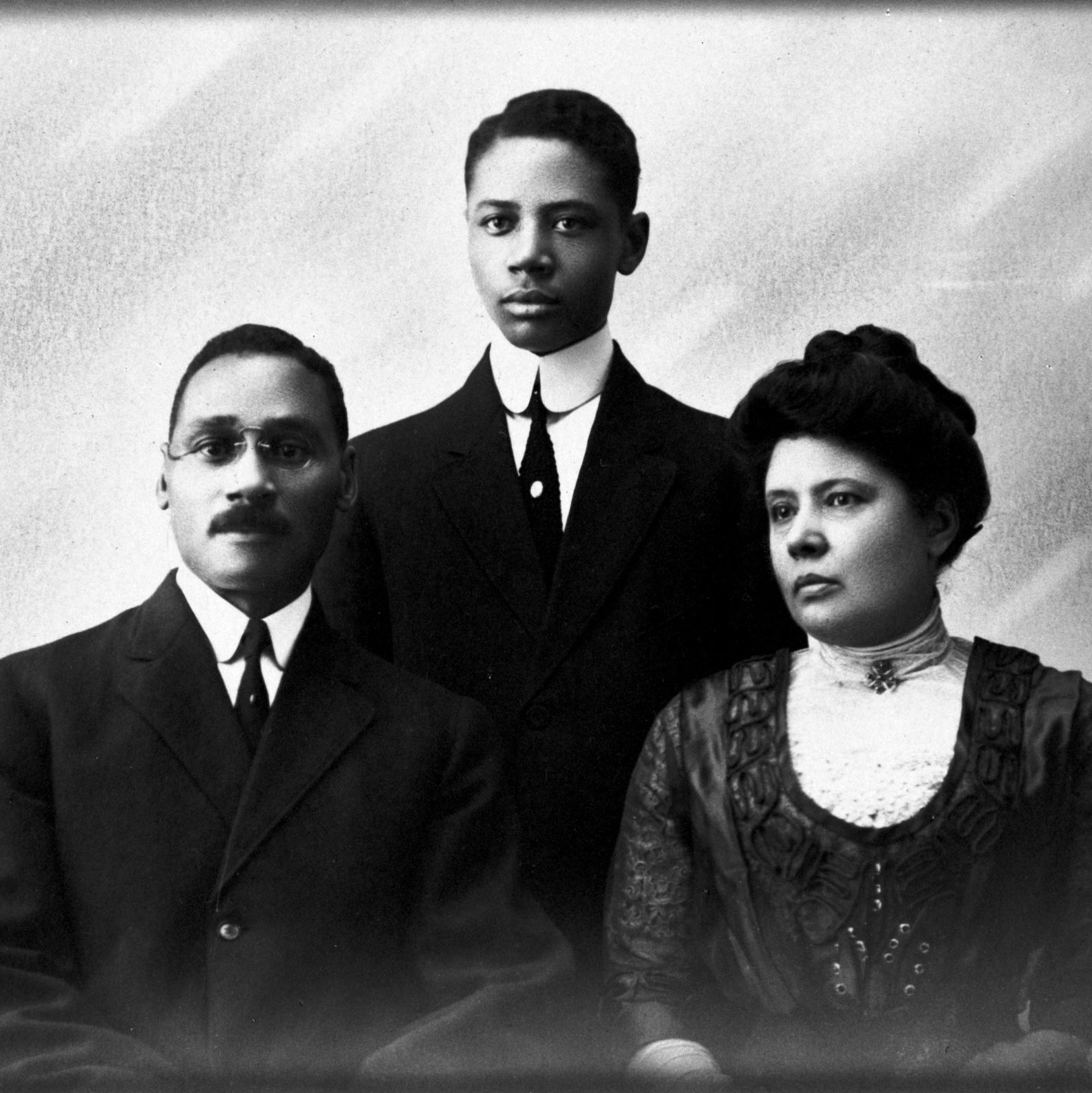 A teenage Charles Houston poses for a portrait with his parents, seated in front of him. His father, William Le Pre Houston, sits to the left, while his mother, Mary Hamilton Houston, is seated on the right.