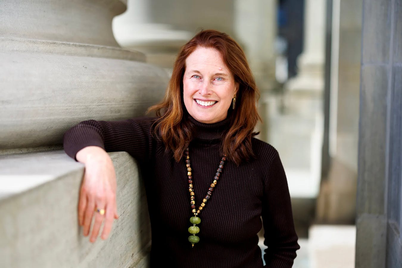 A smiling woman in a black turtleneck and long necklace rests her right arm against the ledge on a large stone column.