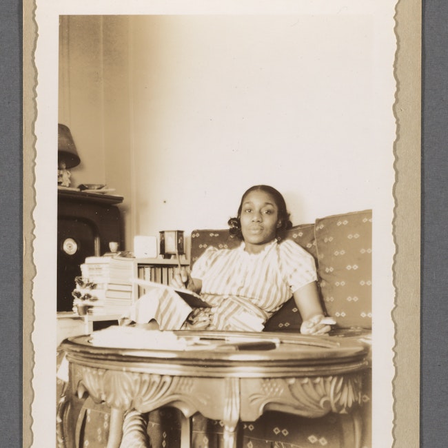 Dorothy West in her New York City apartment. West is seated on a couch, holding a pad of paper on her lap