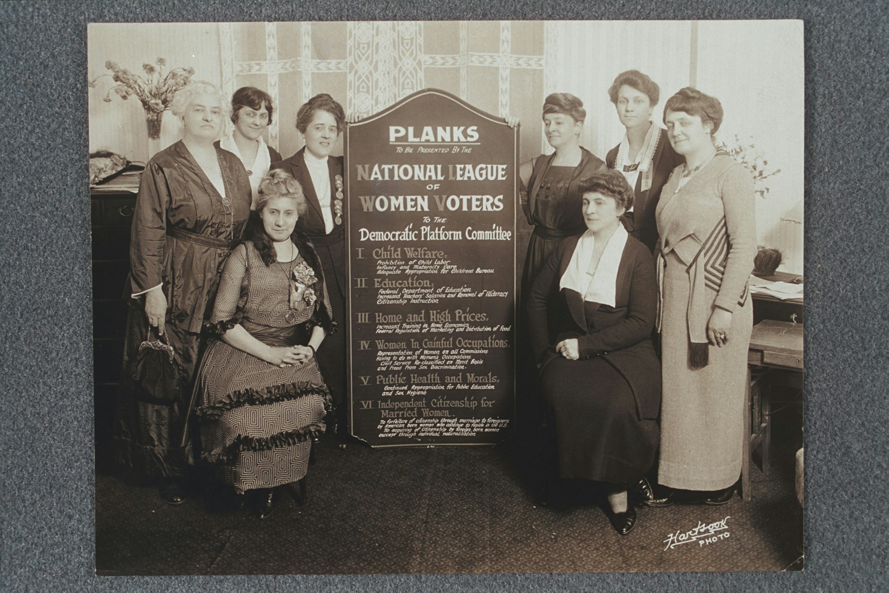 Group portrait of eight women holding a sign listing the planks to be presented by the NLWV to the Democratic Platform Committee. Pattie Ruffner Jacobs (left) and Maud Wood Park (right) are holding the sign