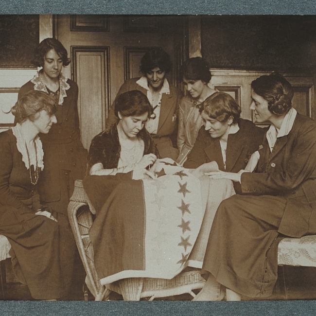 Photograph of Alice Paul sewing stars on the suffrage flag as others look on