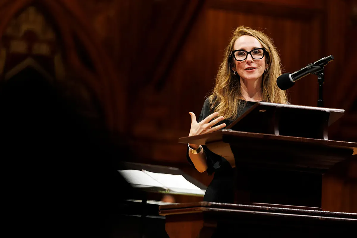 A woman wearing glasses speaks at a podium in dramatic indoor lighting.