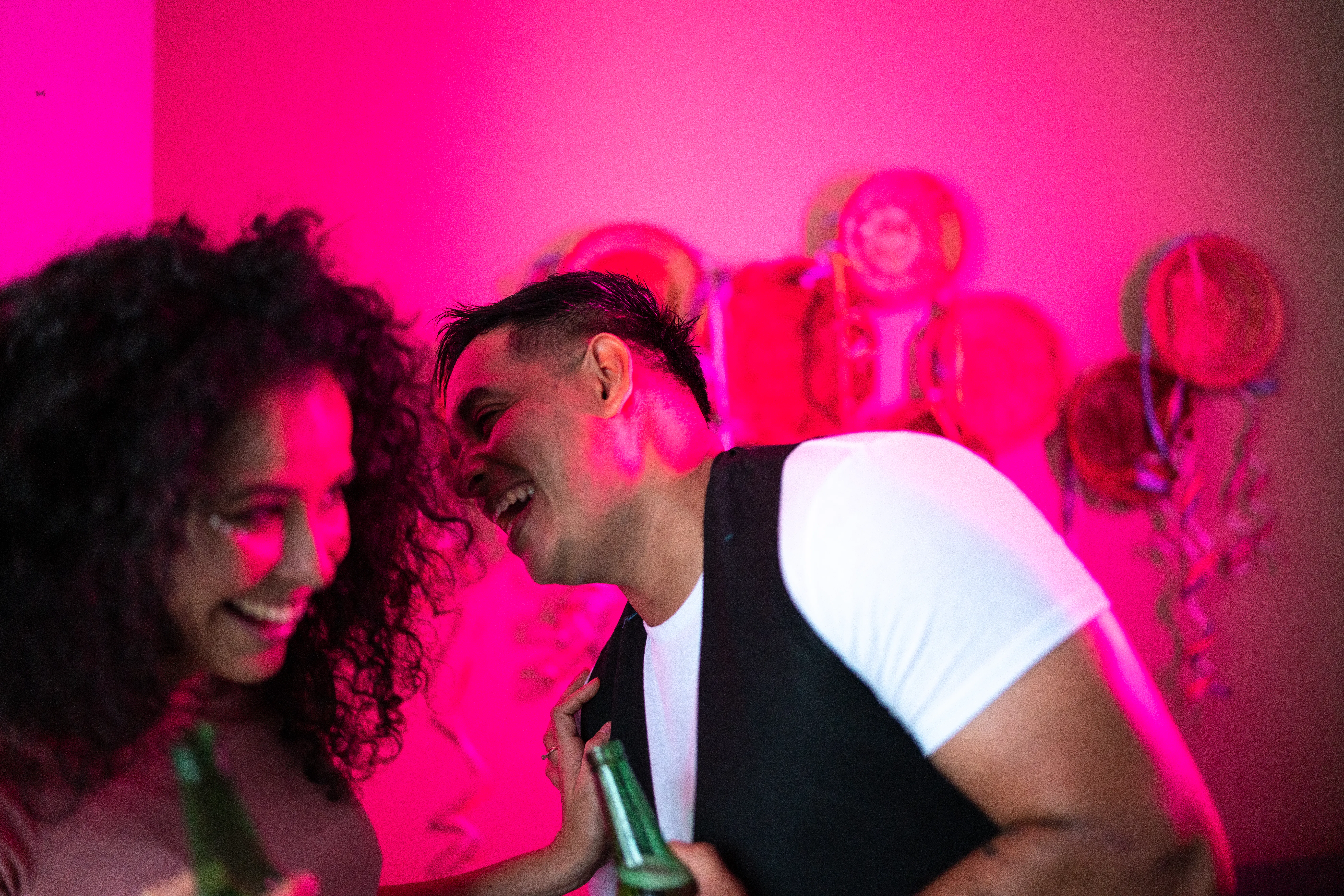A smiling couple holding beer bottles talk at a party. The woman holds her hand up to the man's shoulder, keeping him at arm's length.