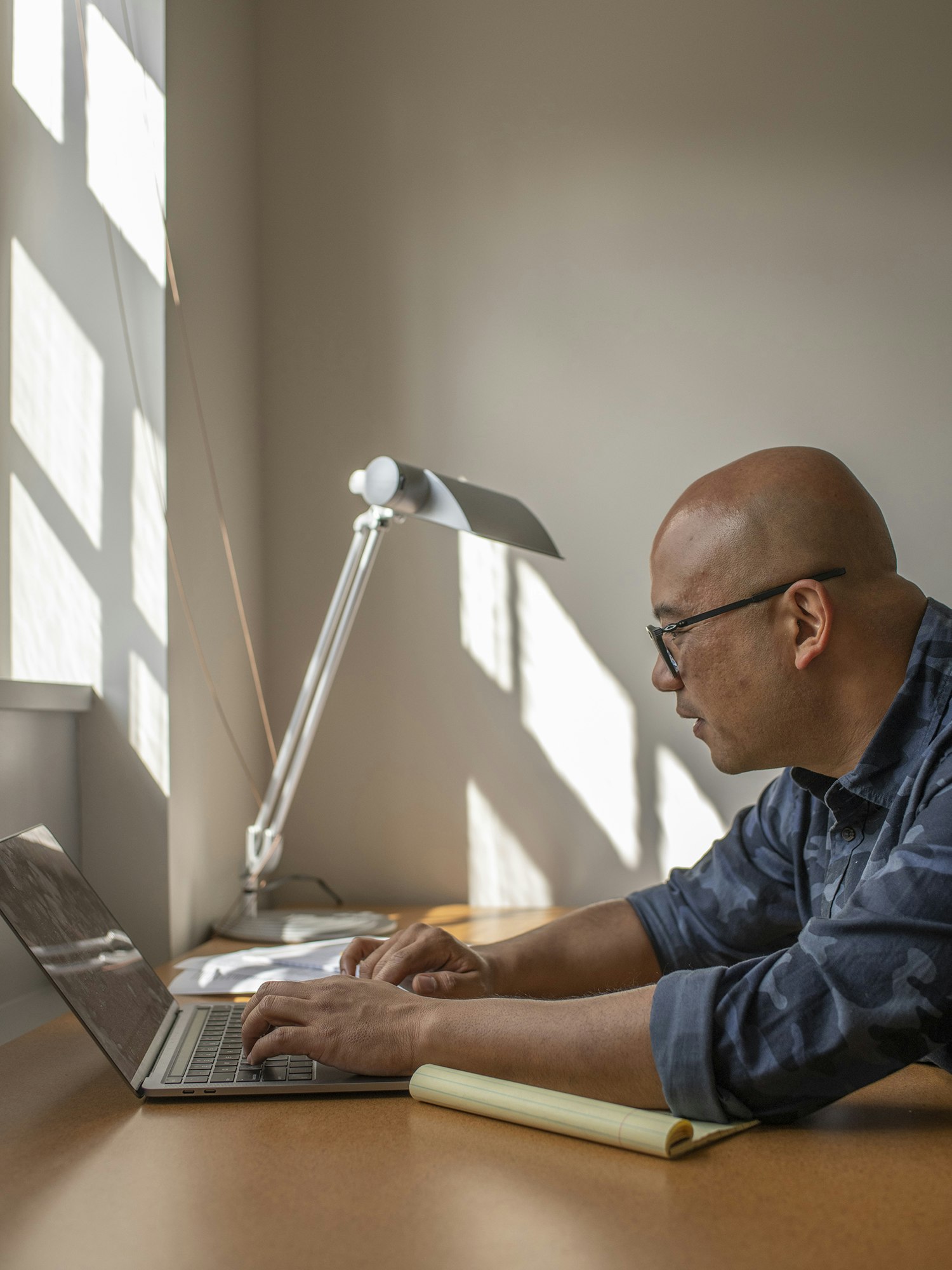A man, in profile, types on a laptop as the sun streams in the window.