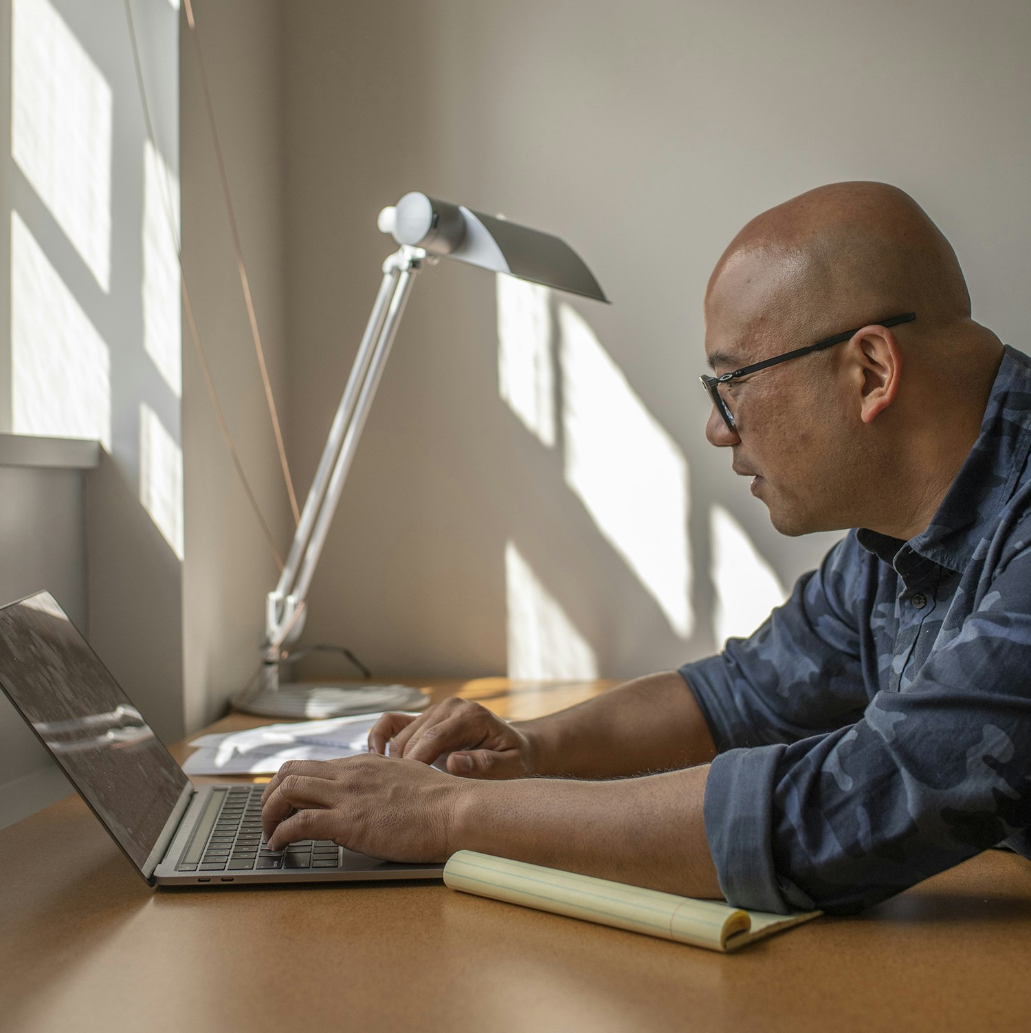 A man, in profile, types on a laptop as the sun streams in the window.