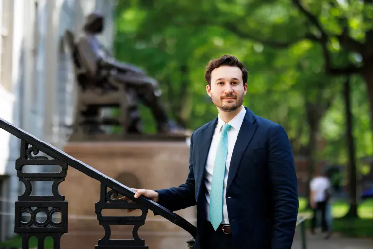A young man wearing a suit and an azure tie poses outdoors in front of the John Harvard statue.