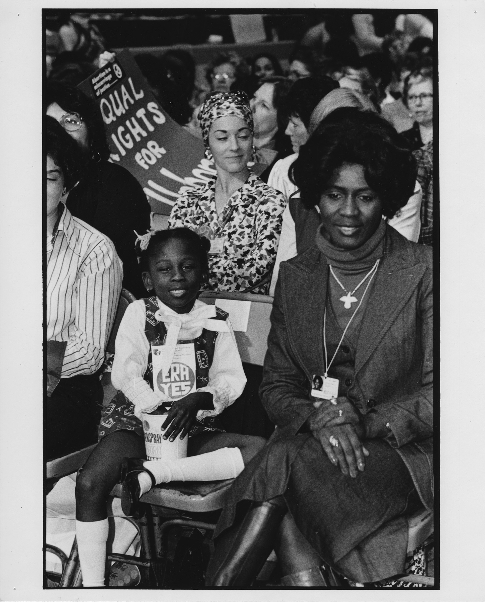 Black and white photograph of a Black woman and her daughter, both smiling, sitting among a conference hall crowd of women.