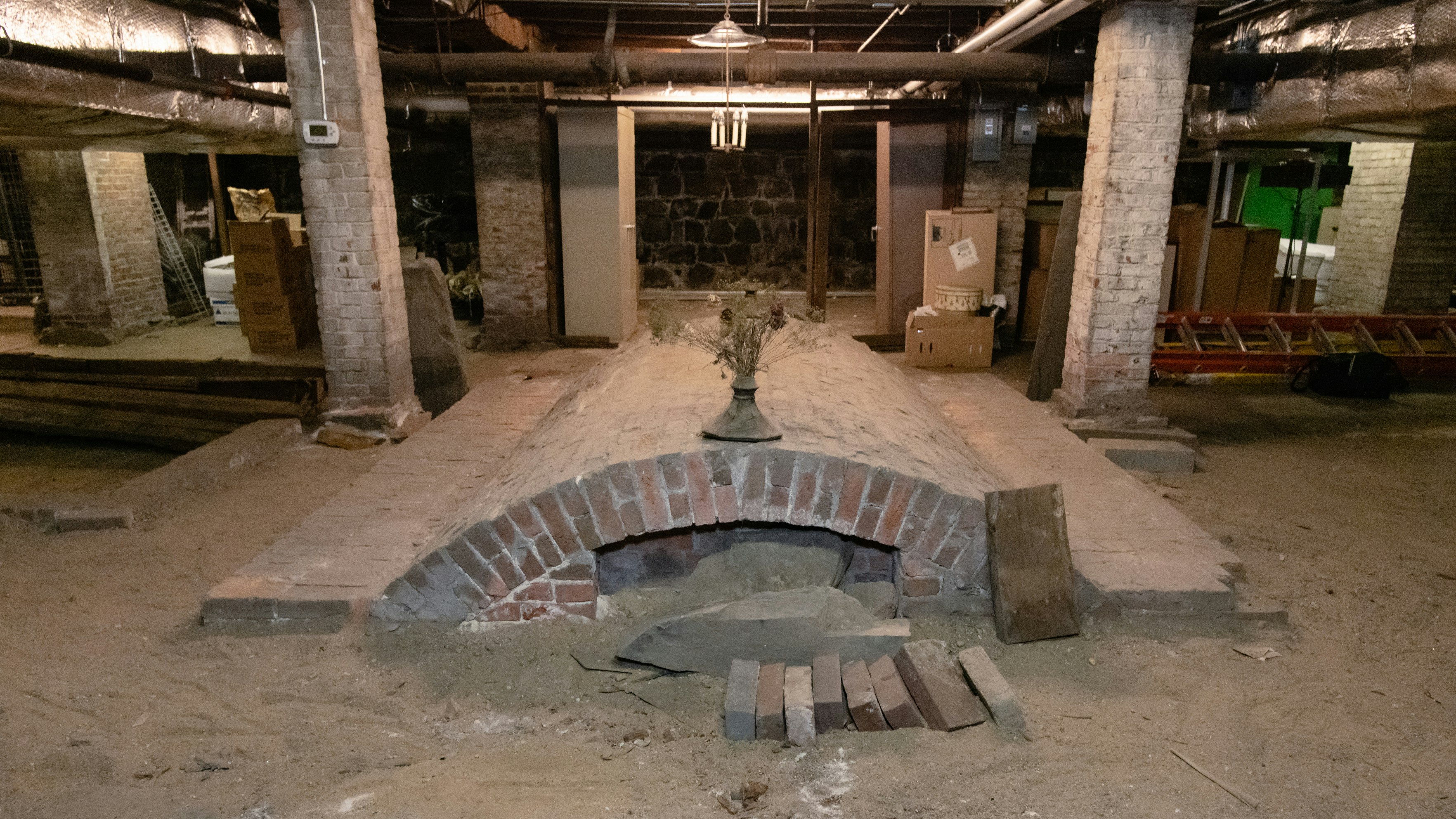 An unfinished, dirt-floored basement with cardboard boxes and several ladders scattered about. At the center, a curved brick structure that contains a tomb.