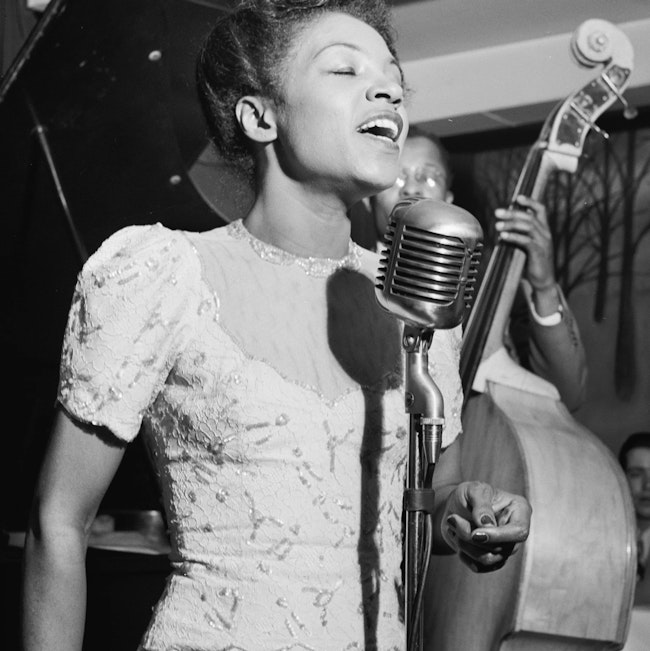 A black-and-white photo of a Black woman, eyes closed and hands at her sides, singing at a vintage microphone; in the background, a man plays stand-up bass.