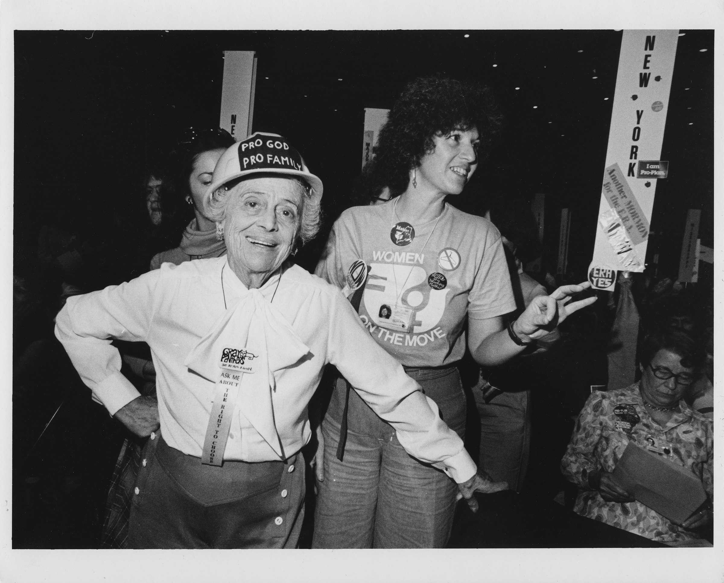 An older woman wearing a hardhat with the words "PRO GOD PRO FAMILY," a Gray Panthers pin, and a ribbon that reads "Ask me about the right to choose" smiles at the camera amid a conference crowd of women.