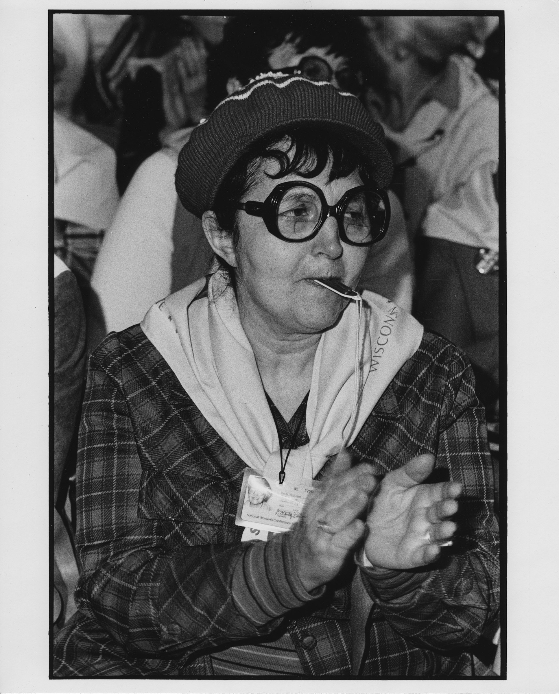 J. Mandy Stellman, wearing bold black round glasses and a jaunty knitted beret, claps while blowing a whistle.
