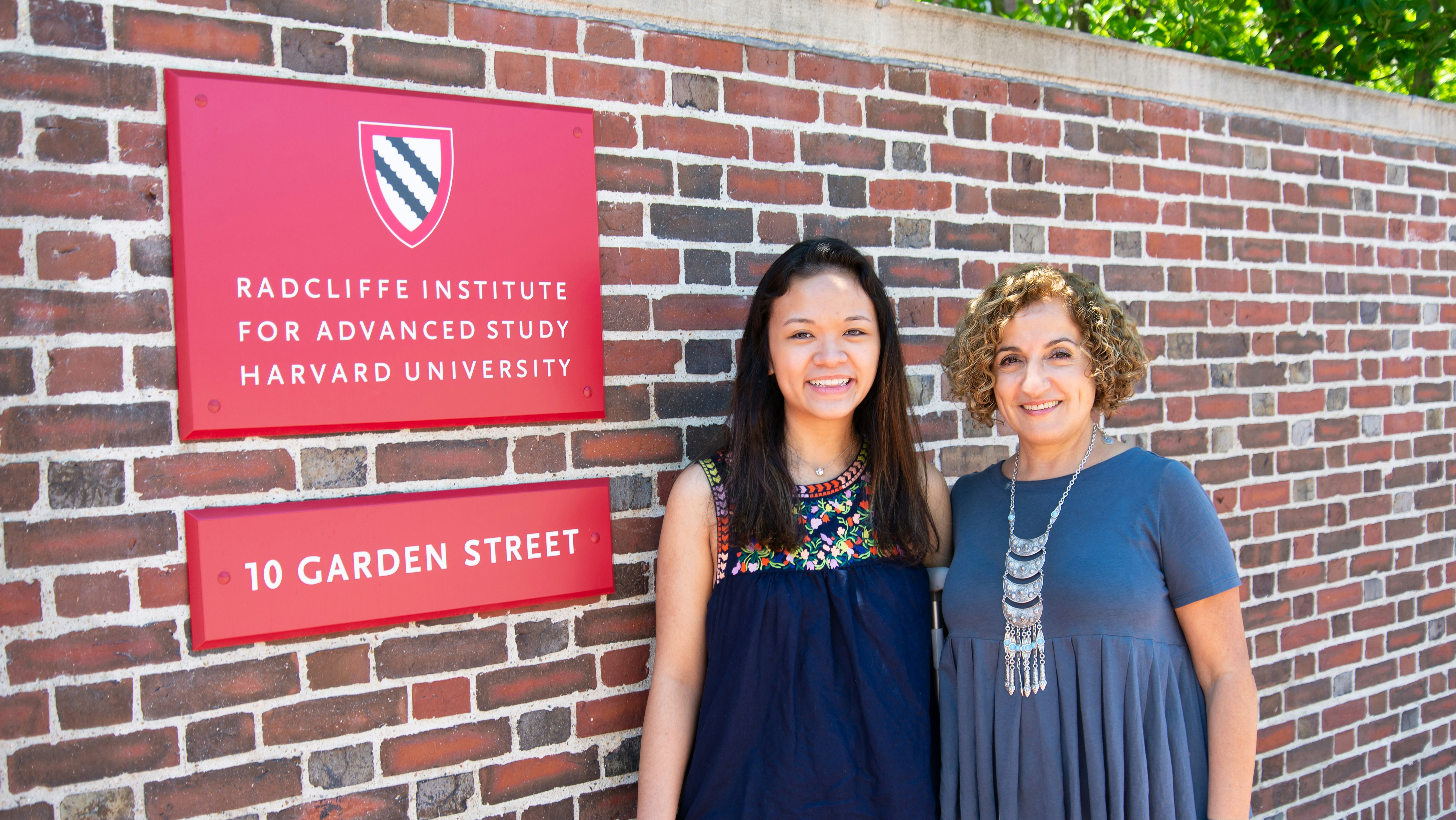 Two women stand next to a red sign for the Radcliffe Institute for Advanced Study posted on a brick wall.