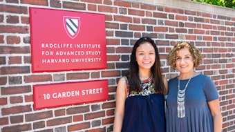 Two women stand next to a red sign for the Radcliffe Institute for Advanced Study posted on a brick wall.