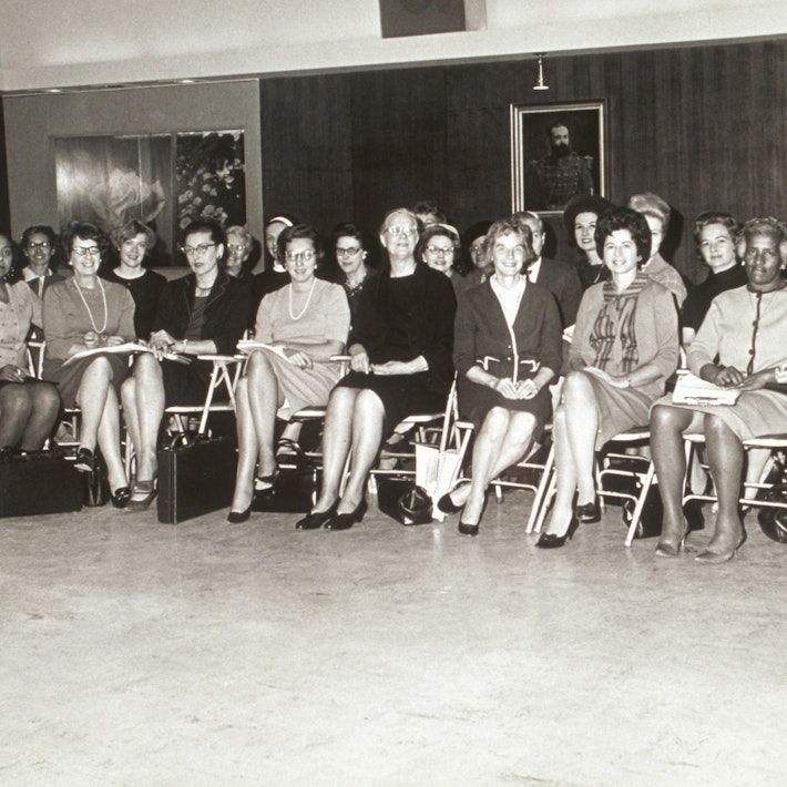 A historical photo of a group of 24 women and one man sitting on folding chairs.