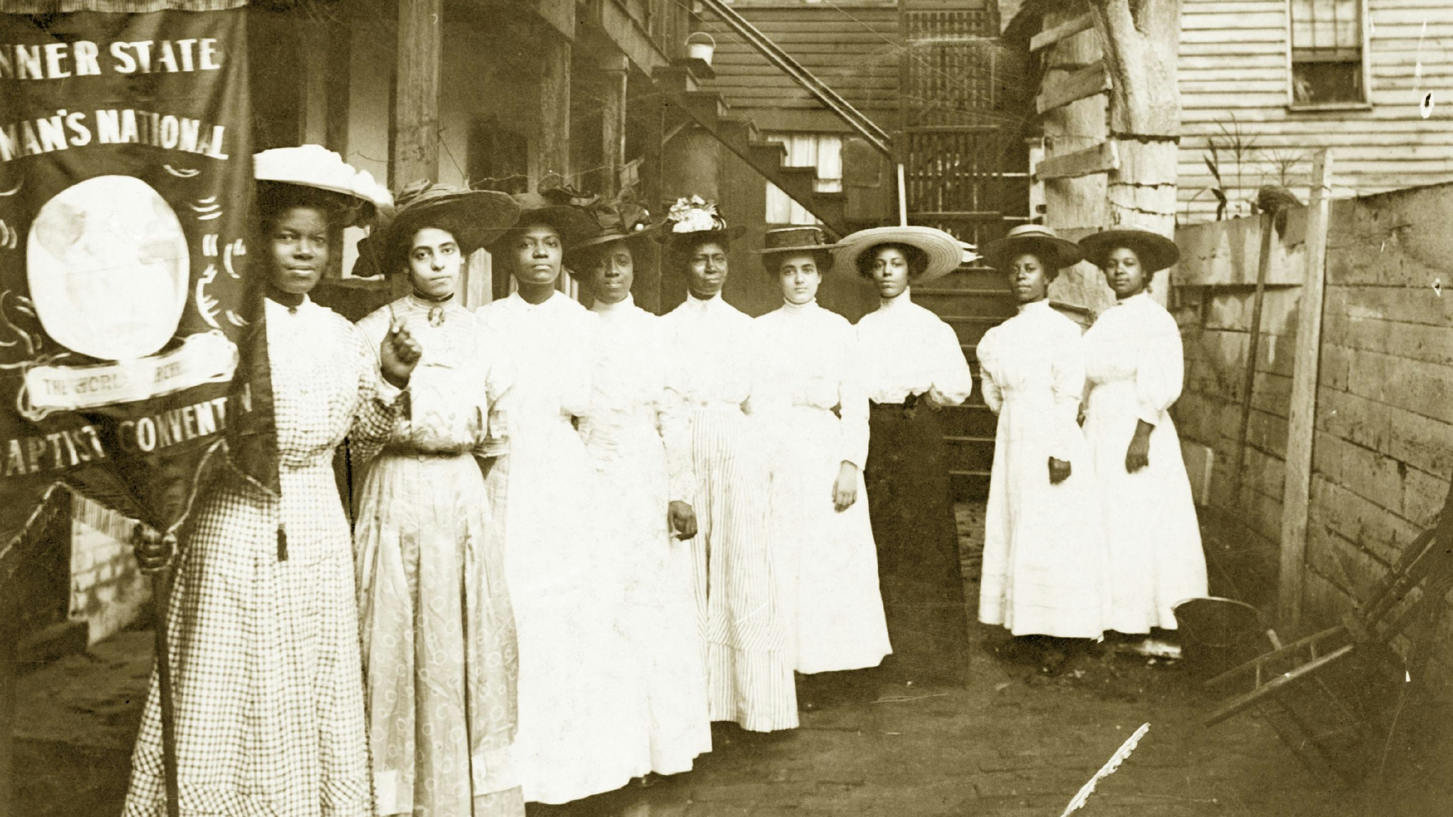 Nine African-American women posed, standing, full length, with Nannie Burroughs holding banner reading, "Banner State Woman's National Baptist Convention"