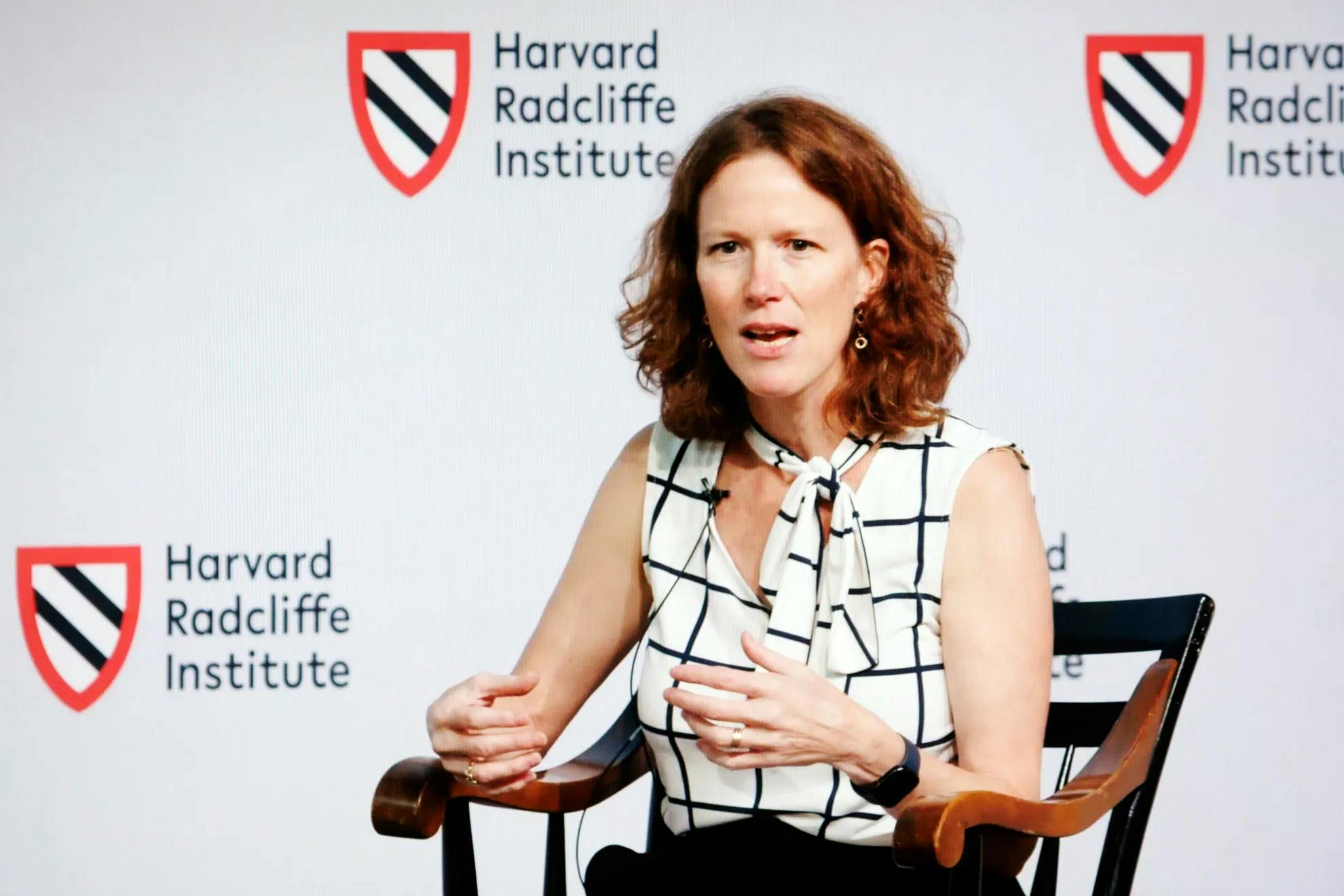 A woman sitting in a captain's chair in front of a Harvard Radcliffe Institute backdrop is caught mid-sentence.