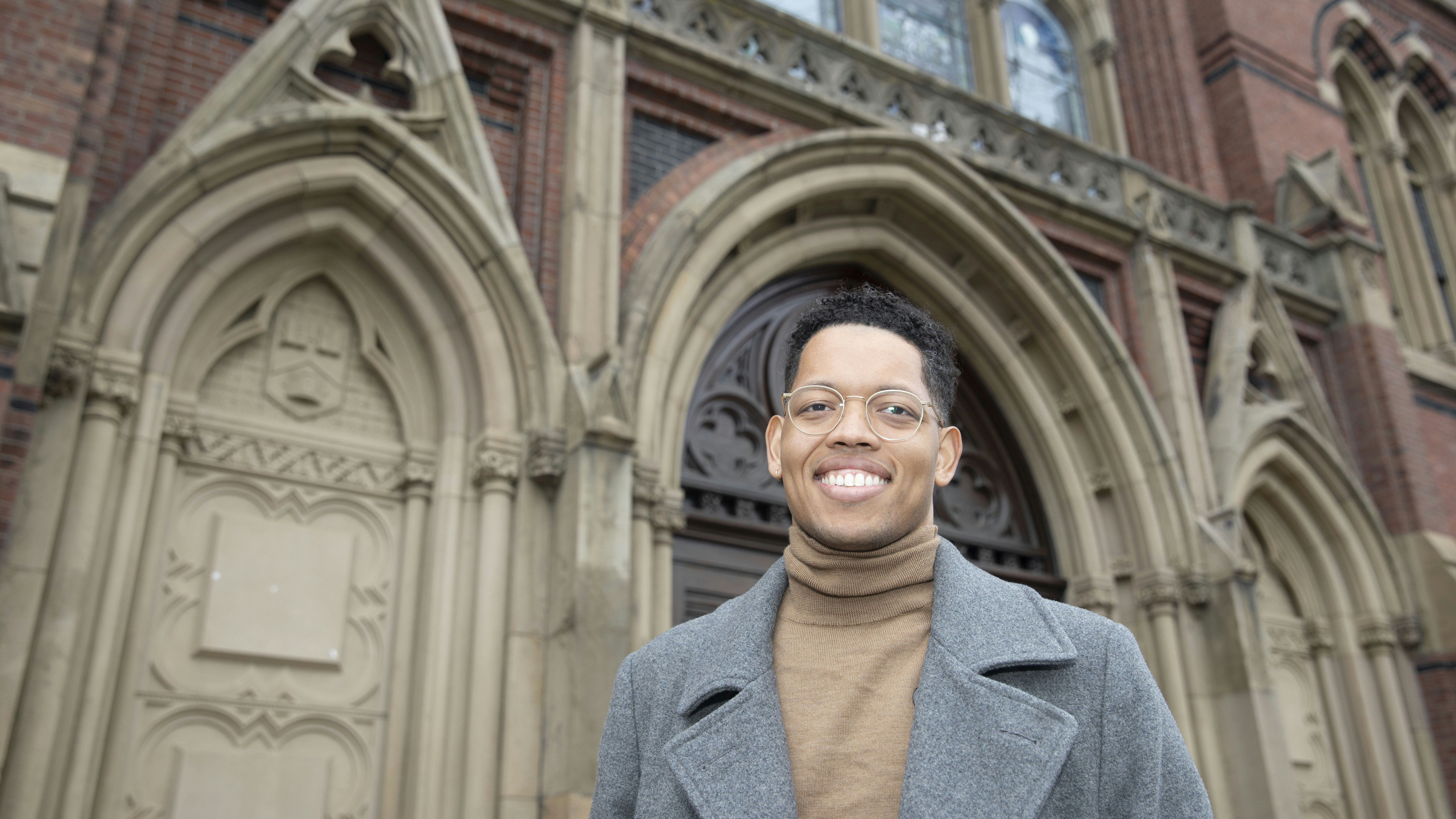 A smiling man in a turtleneck sweater and overcoat stands outside of Memorial Hall