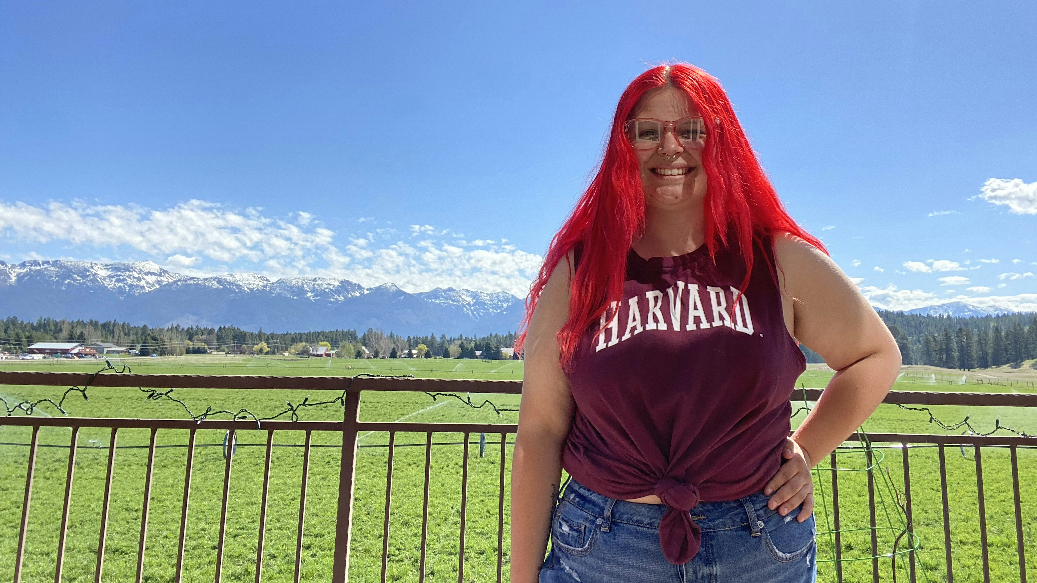 A smiling Liz Hoveland poses outdoors in a Harvard T-shirt. The Rocky Mountains are visible far behind her.