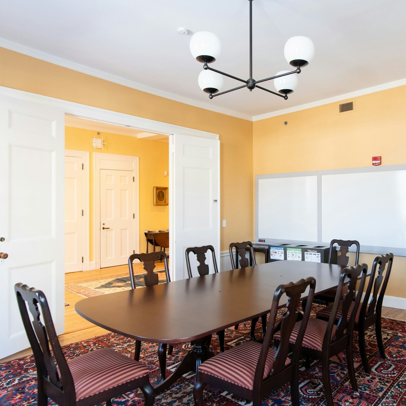 Another angle of dark brown wood conference table with 8 seats lined around. Entrance is in view, with two white doors open, and 3 white boards mounted to the wall behind the table. Walls are yellow.