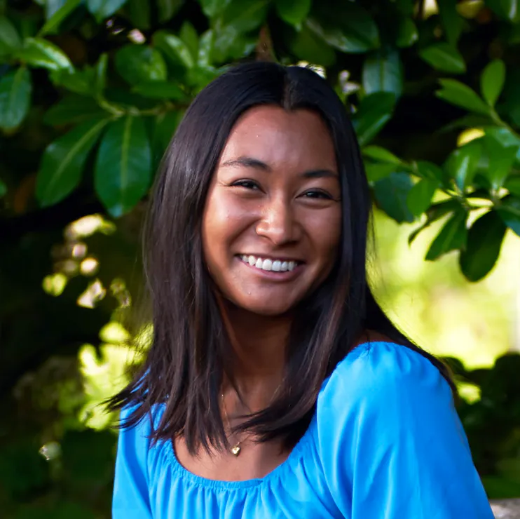 A smiling girl in a bright sky-blue top in a leafy setting.