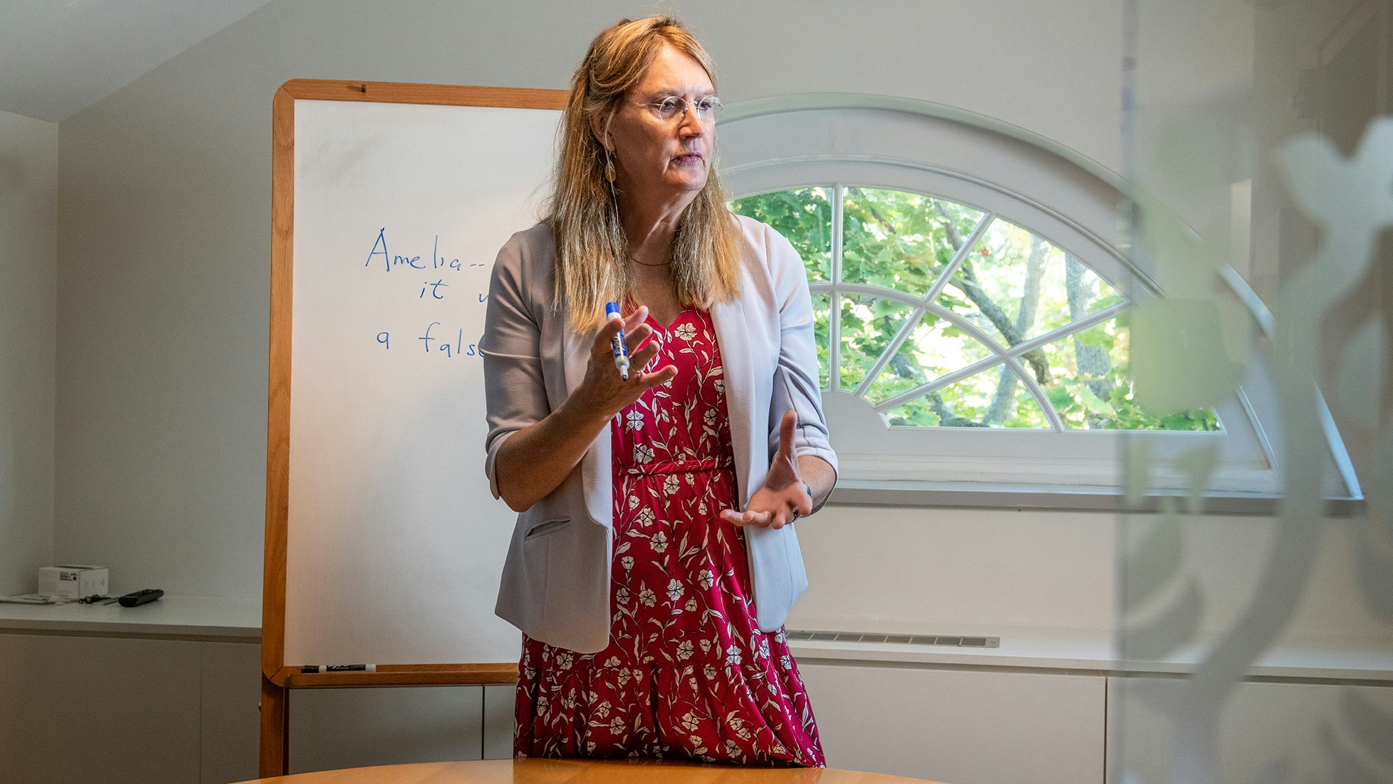 Wearing a serious expression, a bespectacled woman in a silver gray blazer and red patterned dress stands behind a round table in a conference room. Dry-erase marker in hand, she stands in front of a whiteboard, on which the name "Amelia" may be seen alongside other, less legible words.