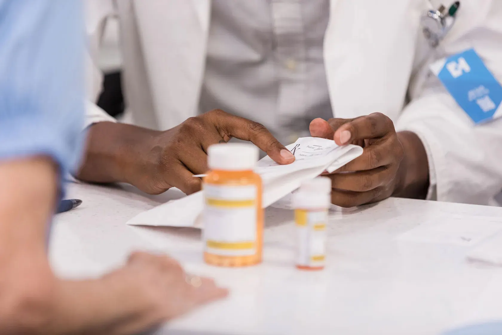 Close-up photo of arms of two people sitting at a table, pill bottles between thm. The person in the background holds a prescription pad and wears a lab coat.