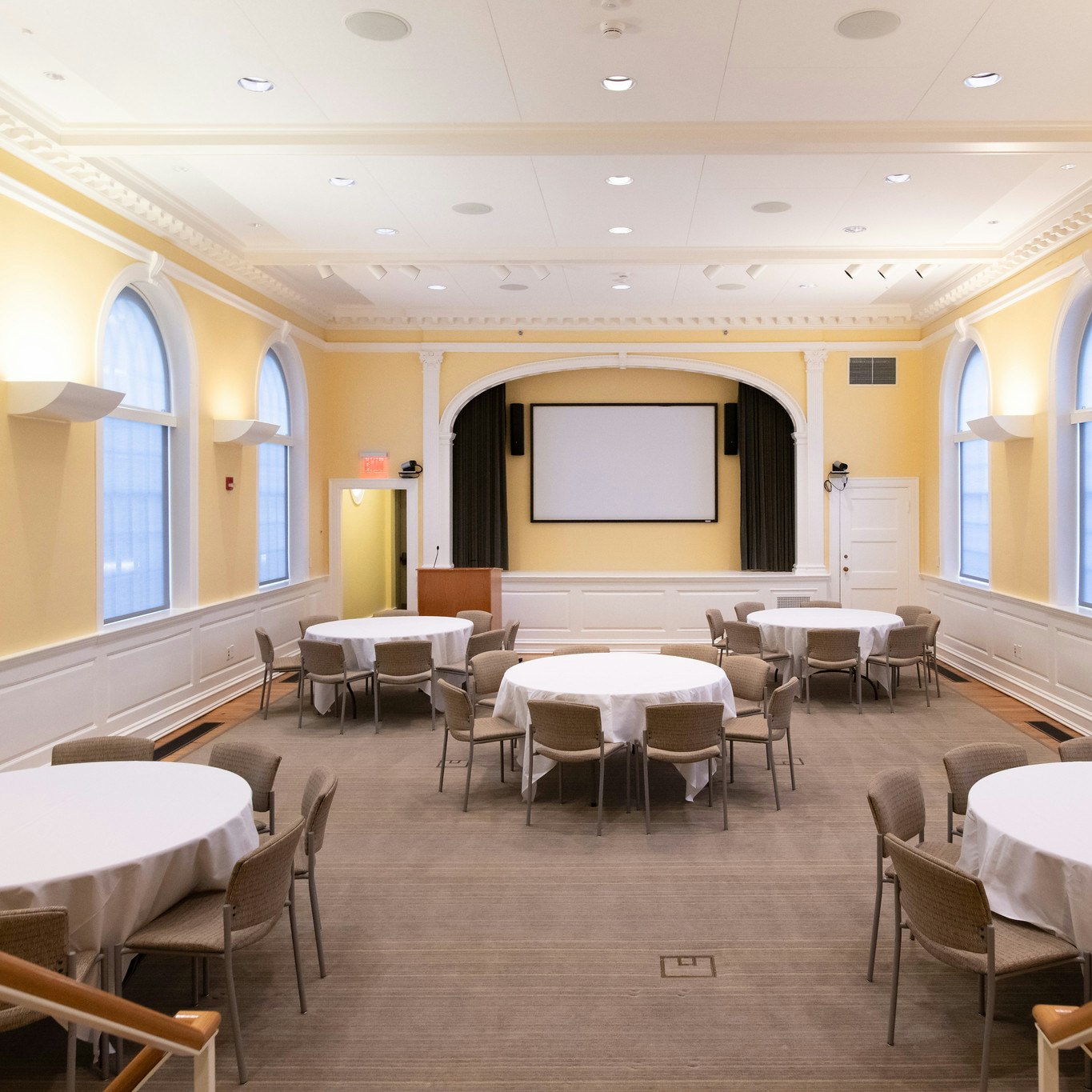 Banquet hall with five circular tables dressed in white table-cloth. Each table seats 8 people. White projector screen is mounted to the wall at the front of the room. View is from the entrance. Walls are yellow.