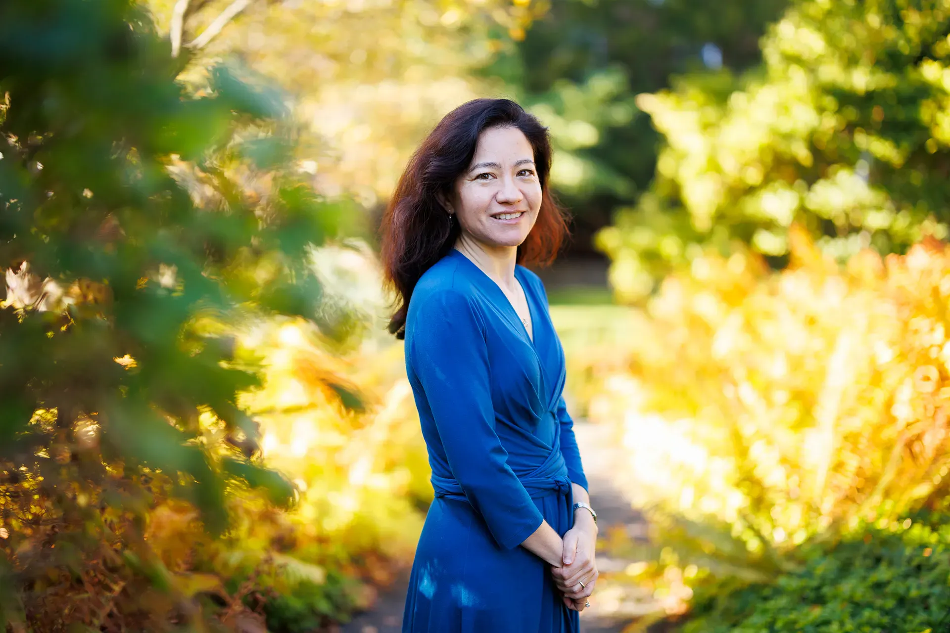 A smiling woman wearing a blue dress seen in three-quarters profile standing outdoors in a very green space.
