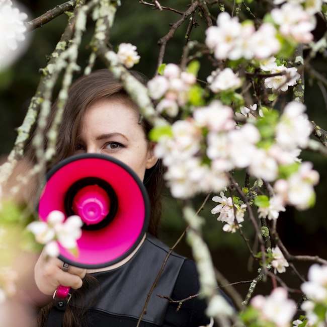 A woman stands among the lilacs, her face partially obscured by a fuchsia megaphone.