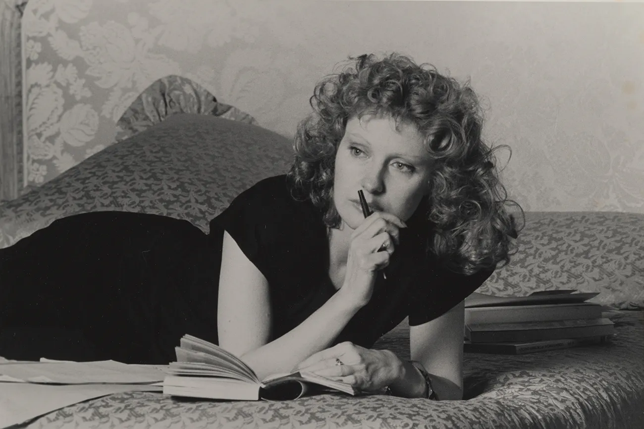 A thoughtful-looking woman lies on her stomach on a bed, propping herself up at the elbows. She holds a notebook open in front of her, a pen in her right hand held up to her mouth.