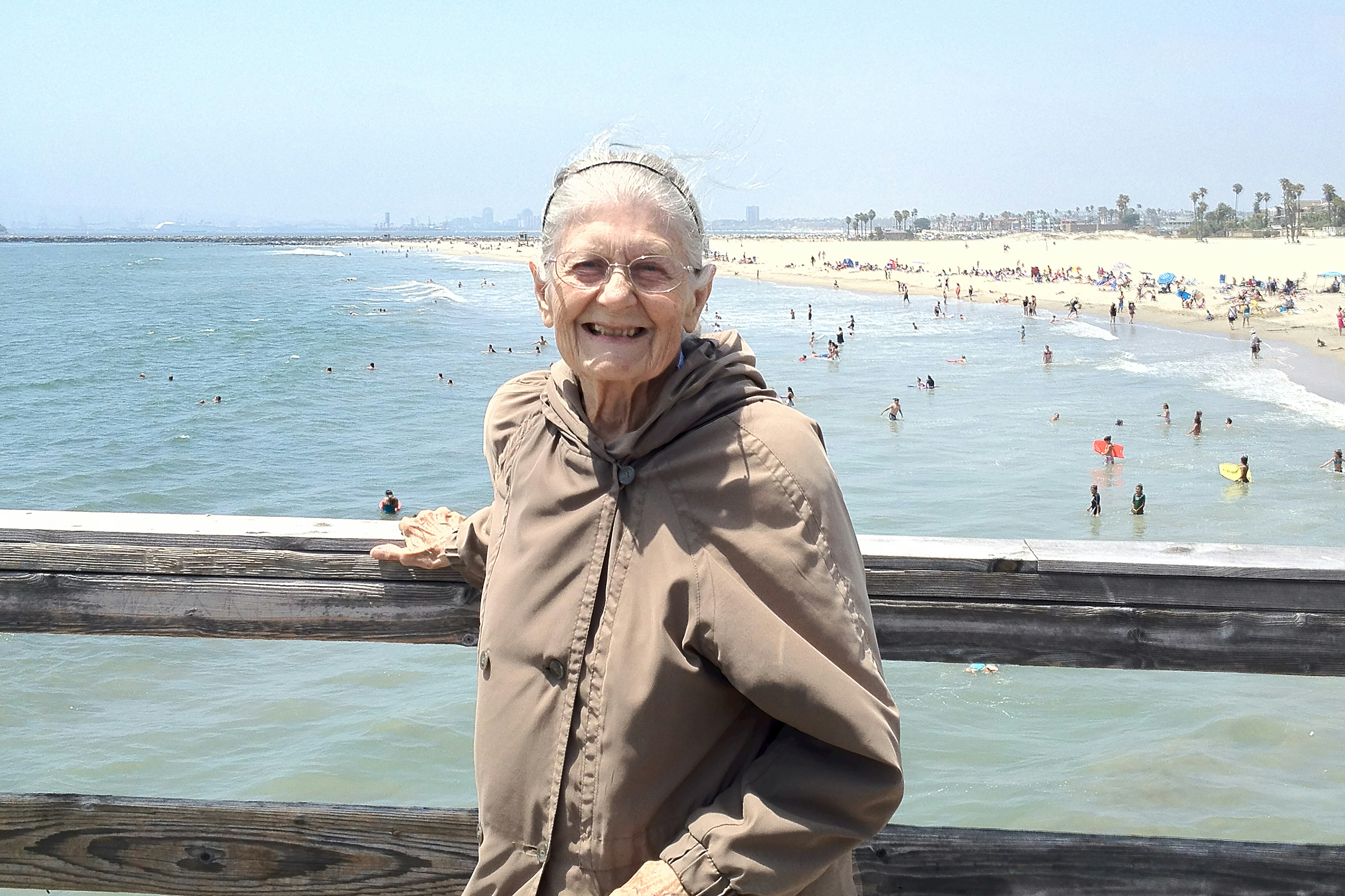 An older woman poses, smiling, at a wooden railing; in the distance behind her, people at the beach.
