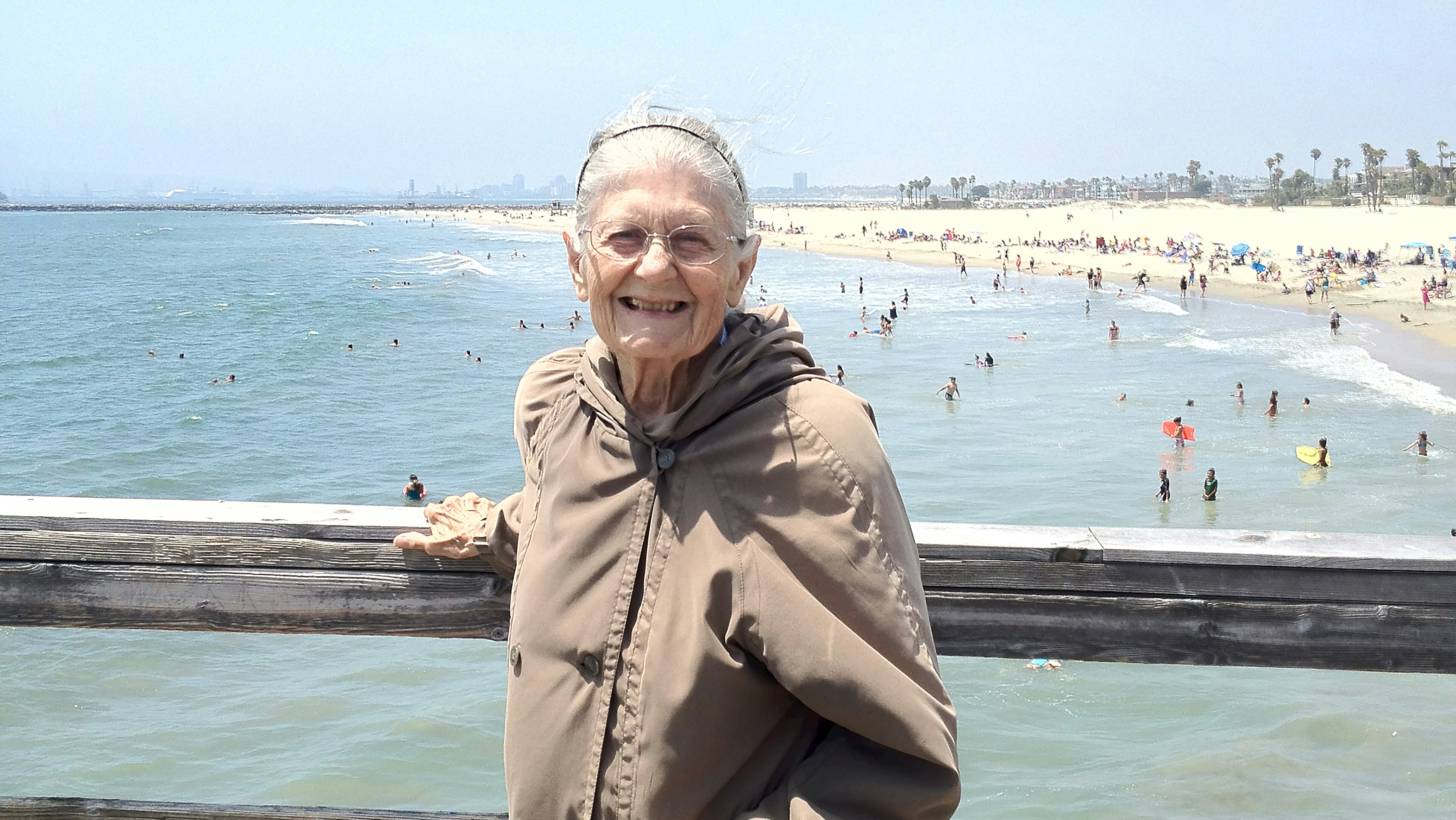 An older woman poses, smiling, at a wooden railing; in the distance behind her, people at the beach.