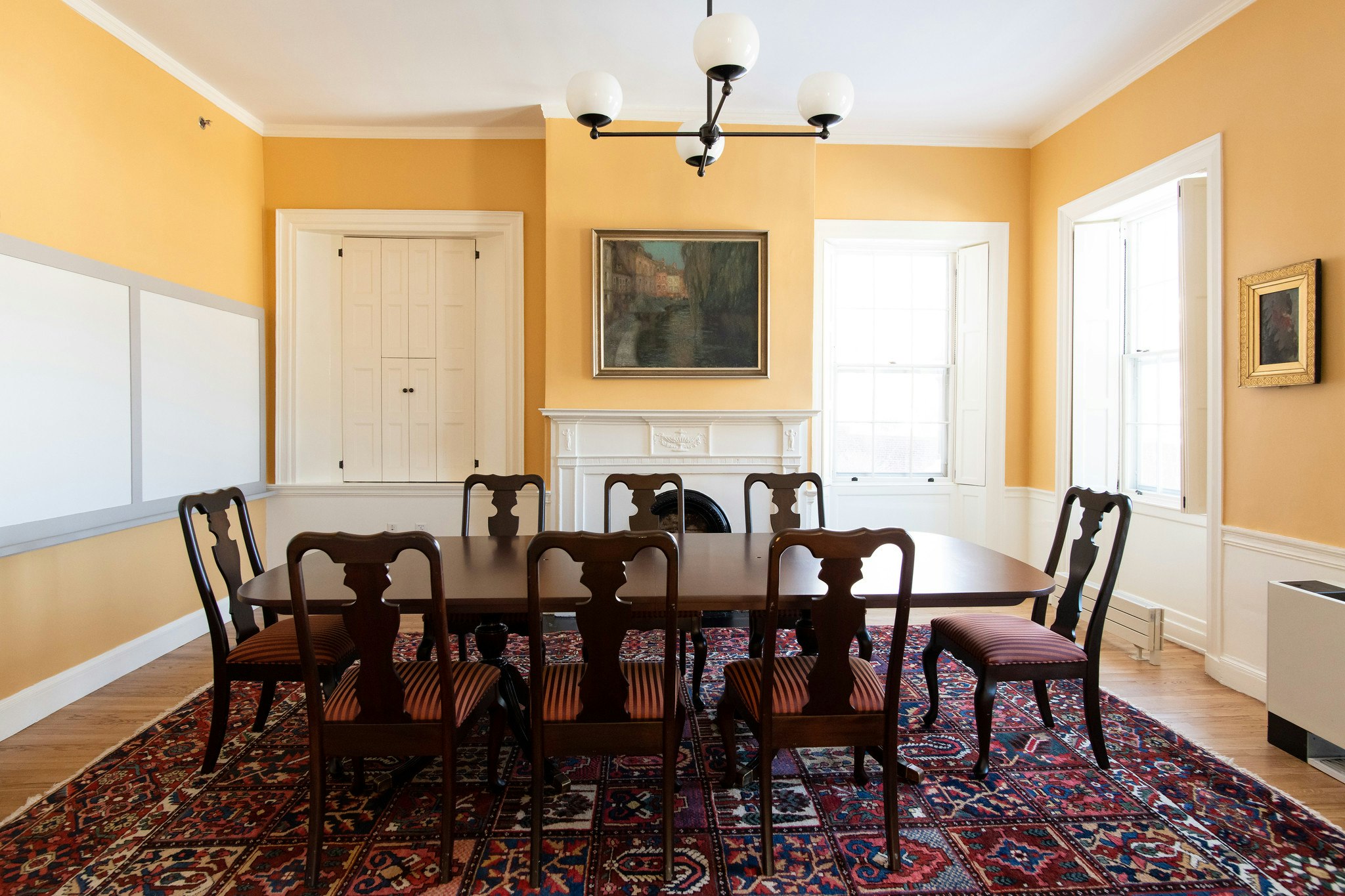 View from the entrance, dark brown wood conference table with 8 seats lined around. Fireplace and painting are on the other side of the room, behind the conference table. Walls are yellow.