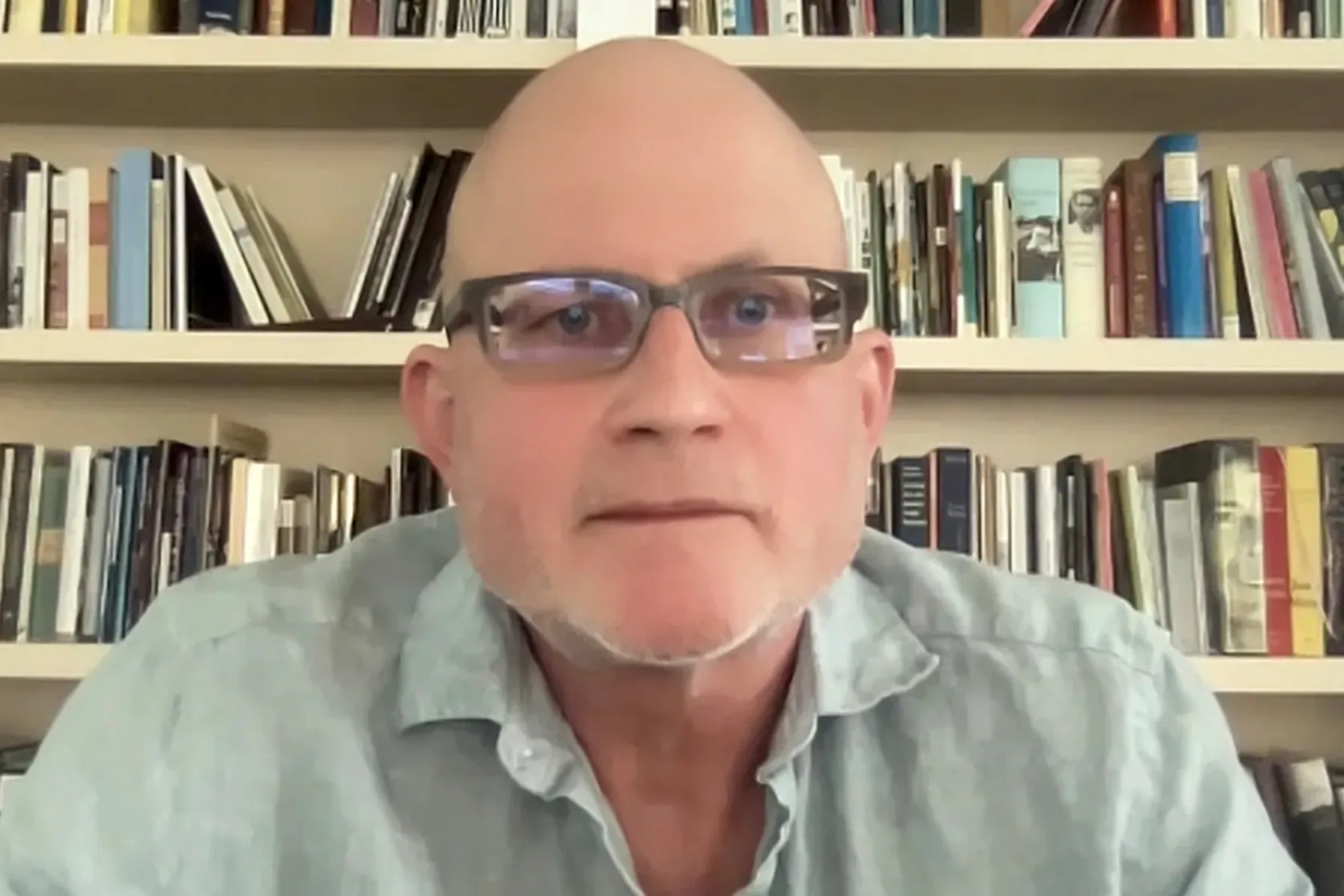 A bald man wearing glasses sitting in front of a filled bookcase stares intently at the viewer.