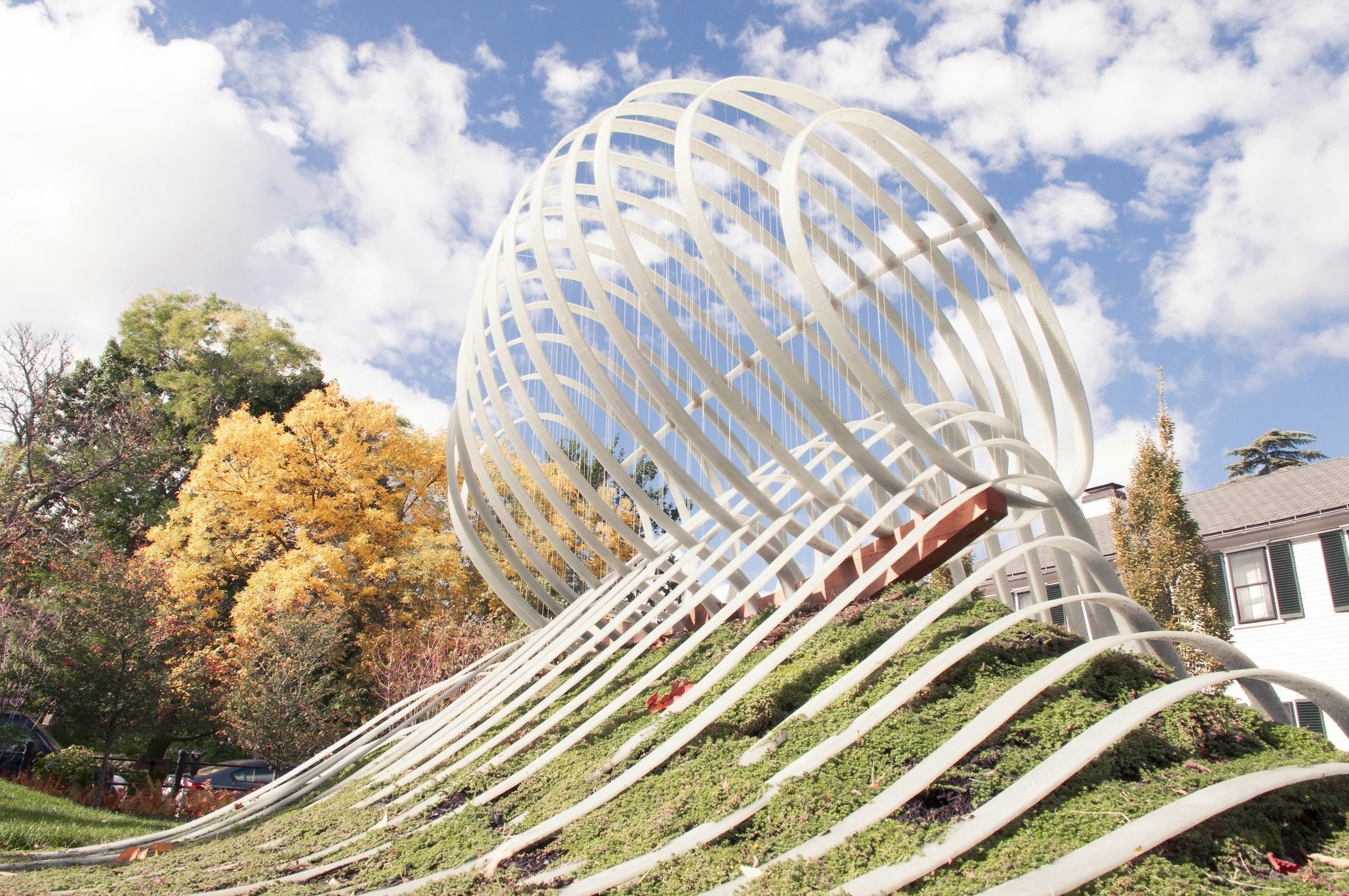 White modern art structure on a grass area. Structure is white, with horizontal grooved lines across the grass, and a sphere of these white lines resting on a small hill. View is closer to the sphere. It is the daytime, the sky is blue with white clouds.