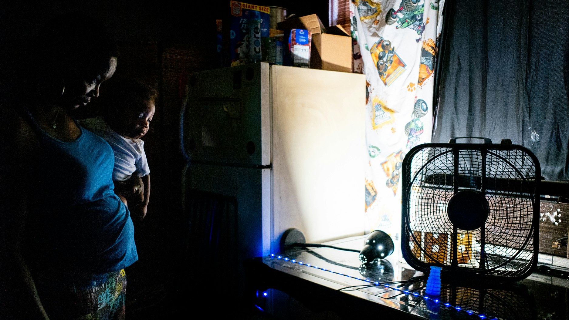 A mother holds her child near a window fan in her darkened home.