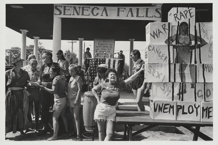 Rally organized by encampment women held in Seneca Falls, New York. A group at left is singing, on right is a display of cartons painted with the image of woman prying open prison bars.