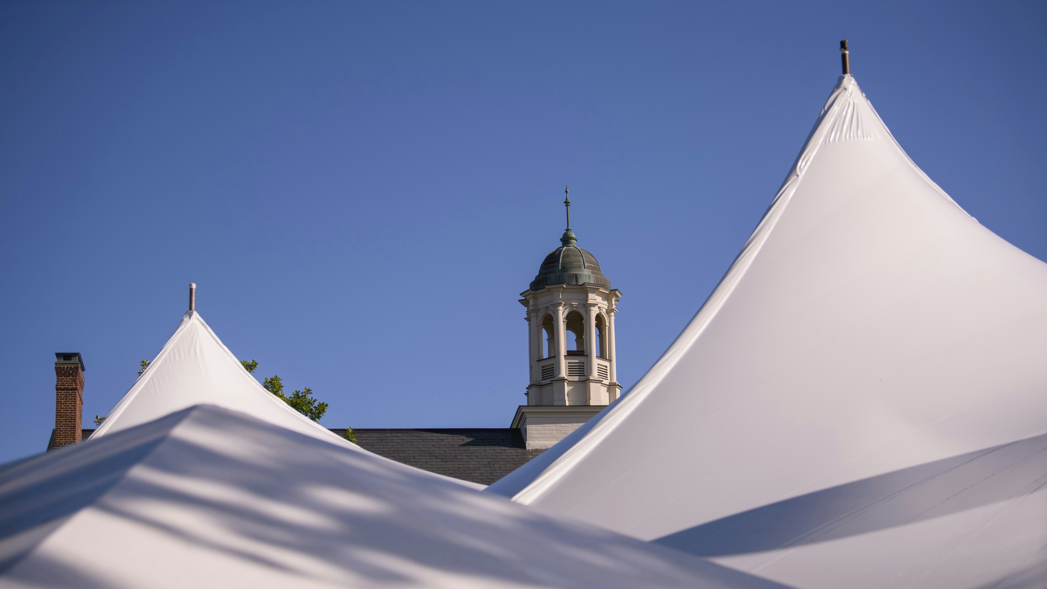 A cloudless blue sky behind the cupola atop the Knafel Center at Harvard Radcliffe Institute. The cupola sits between two large white tent spires set up for events at Radcliffe Day 2023.
