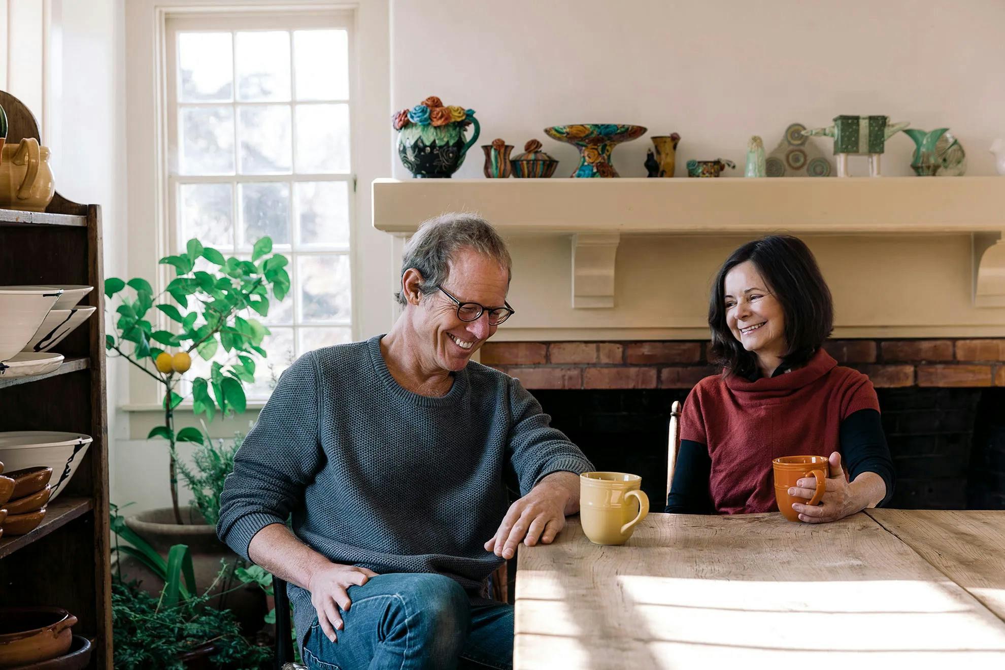 A man and a woman seated at a large wooden ta, laughing over coffee mugs.