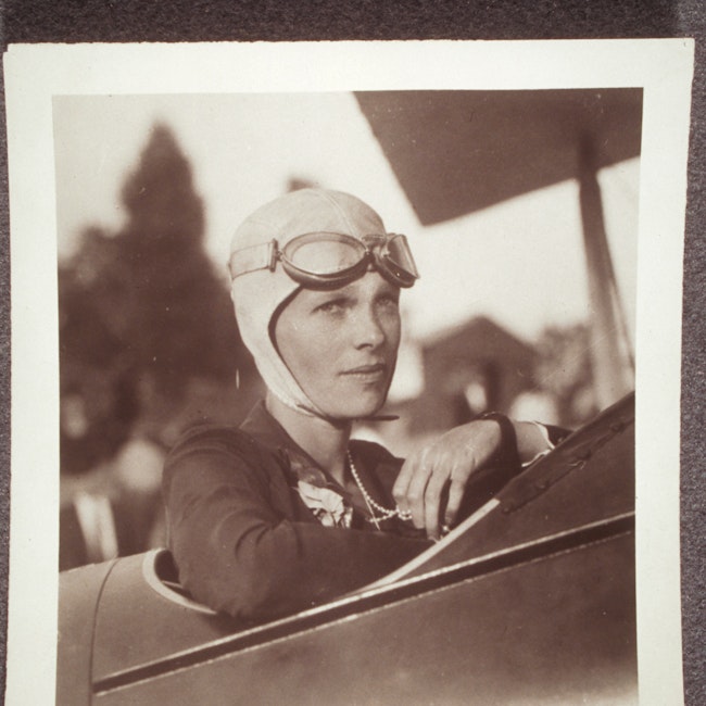 Amelia Earhart, in plane, wearing flight helmet and pearls