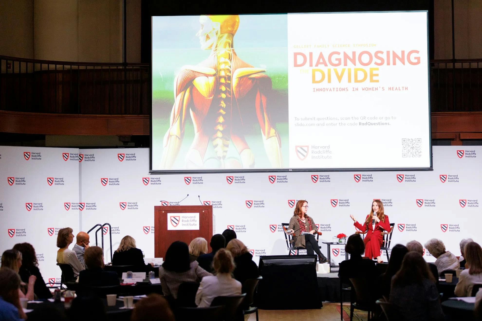 Two women sitting on captain's chairs atop a stage speak in front of a large audience.
