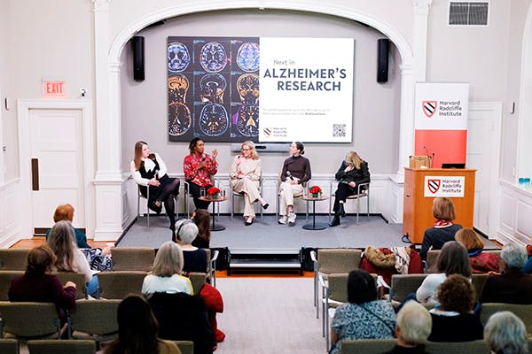 Five women sit on captain's chairs on a small stage in front of a large screen that reads "Next in Alzheimer's Research." There is a seated audience watching them speak.